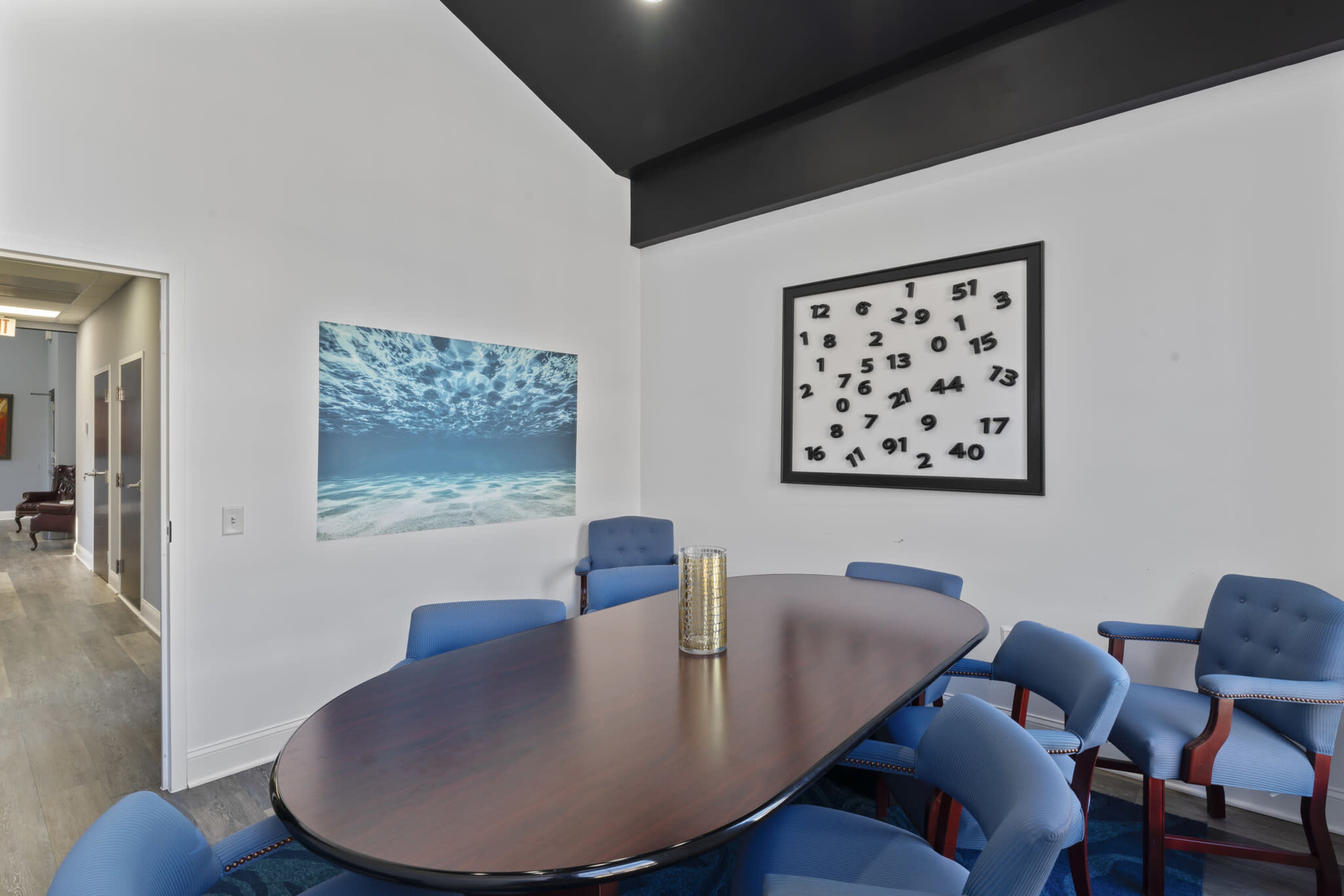 The image shows a modern conference room with a large oval table surrounded by blue chairs, accented by artwork featuring ocean waves and a numerical wall art piece.
