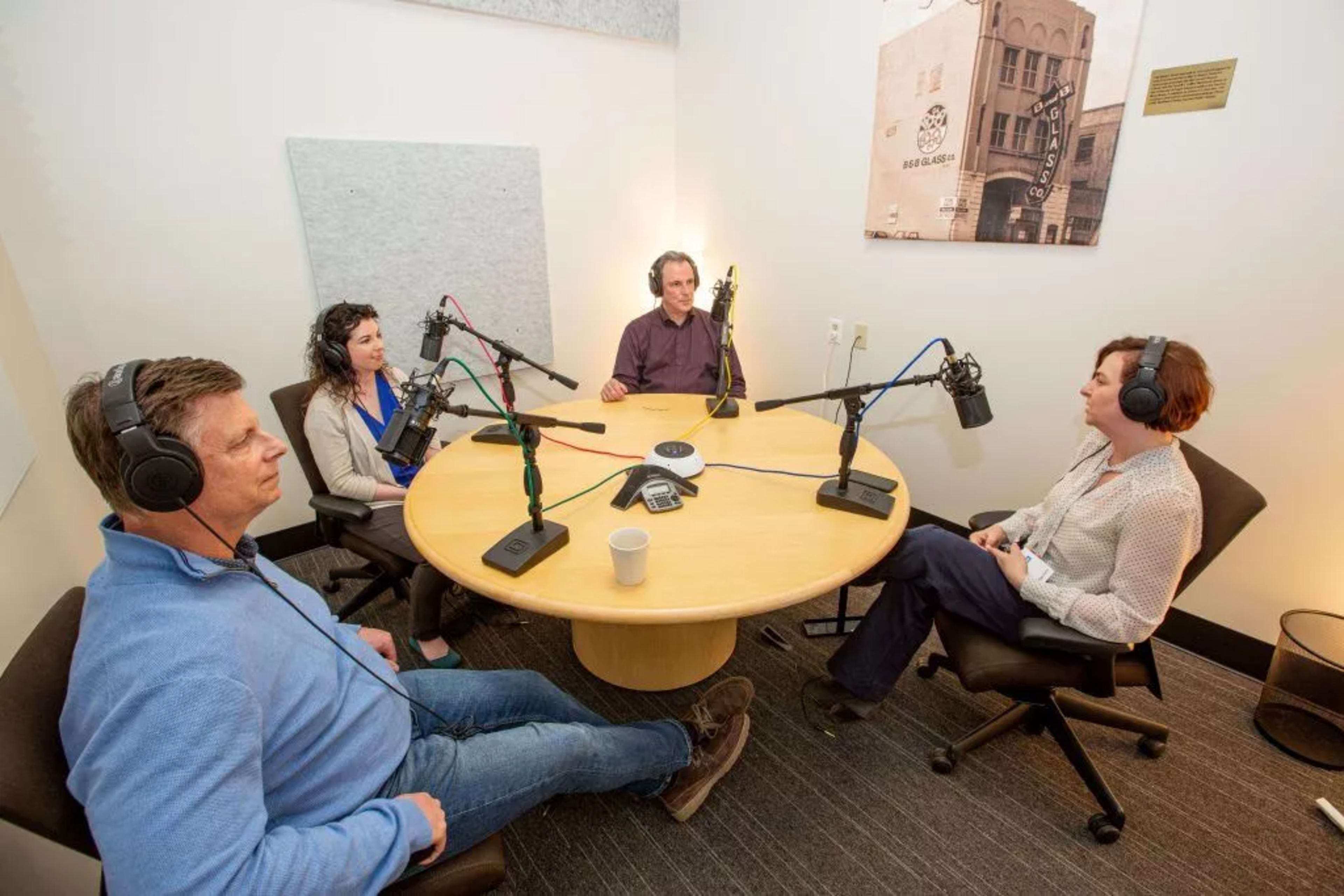 Four people sit around a circular table in a recording studio, each wearing headphones and speaking into microphones.