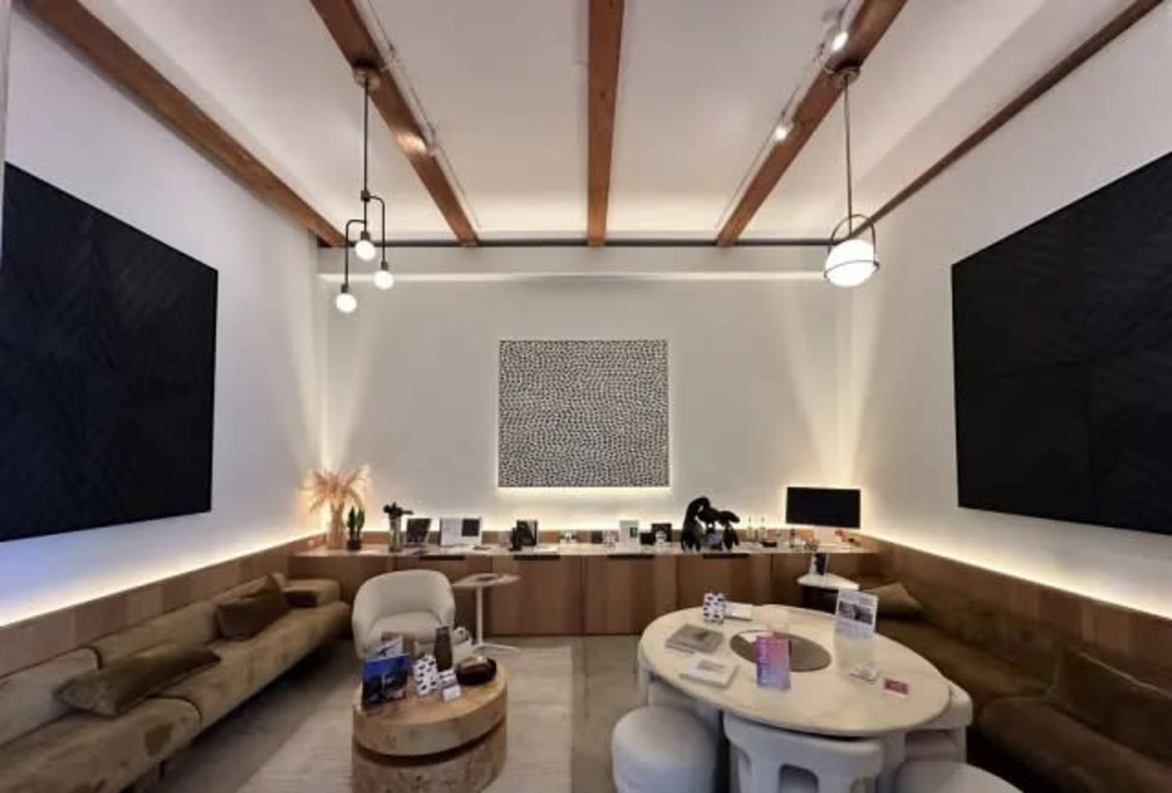 The image shows a modern interior with wooden beams, two large black wall art pieces, a round table in the center, and a seating area along the wall.