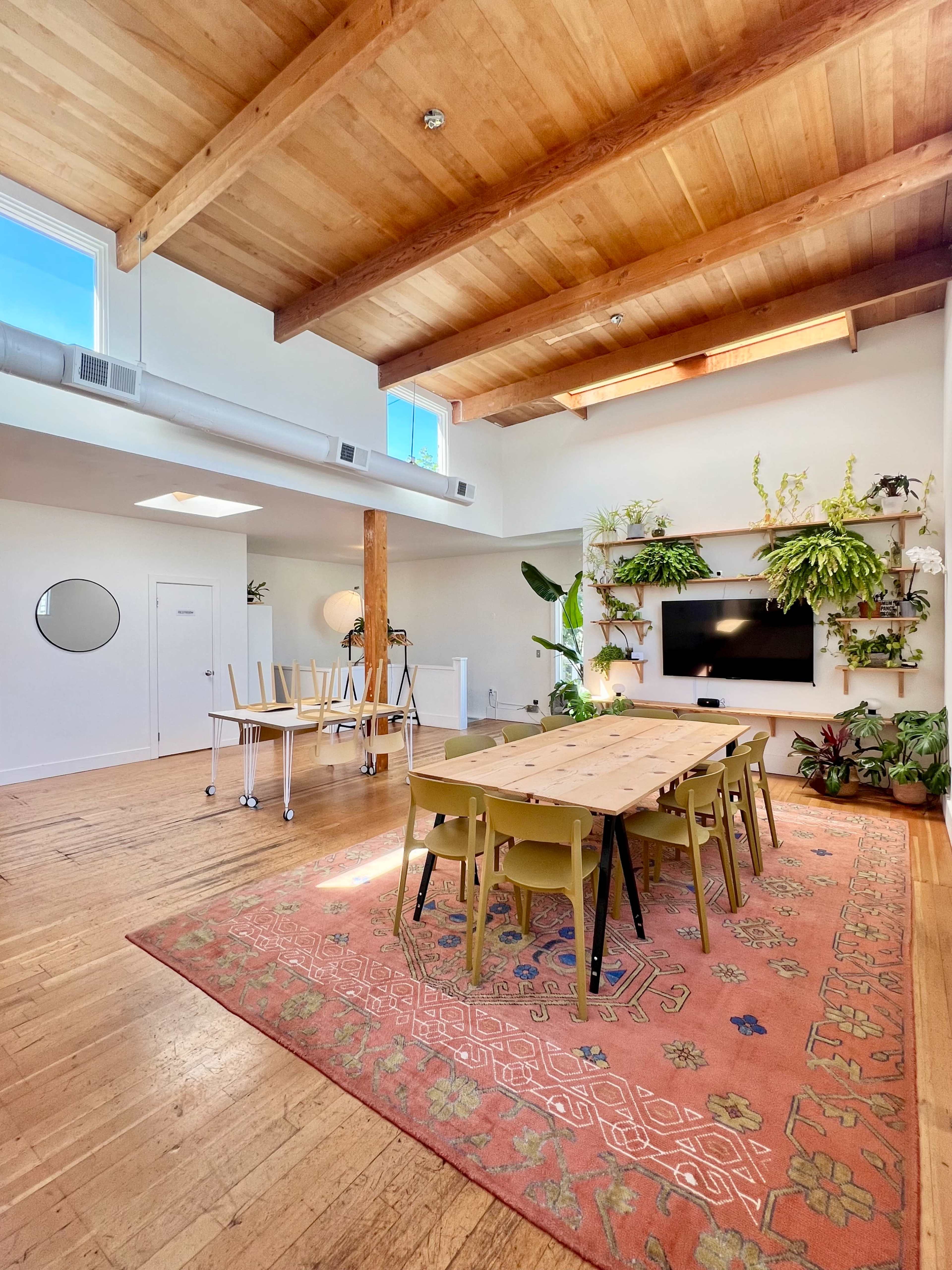 The image shows a spacious room with wooden beams, a long table surrounded by chairs, a television mounted on the wall, and plants arranged on shelves.