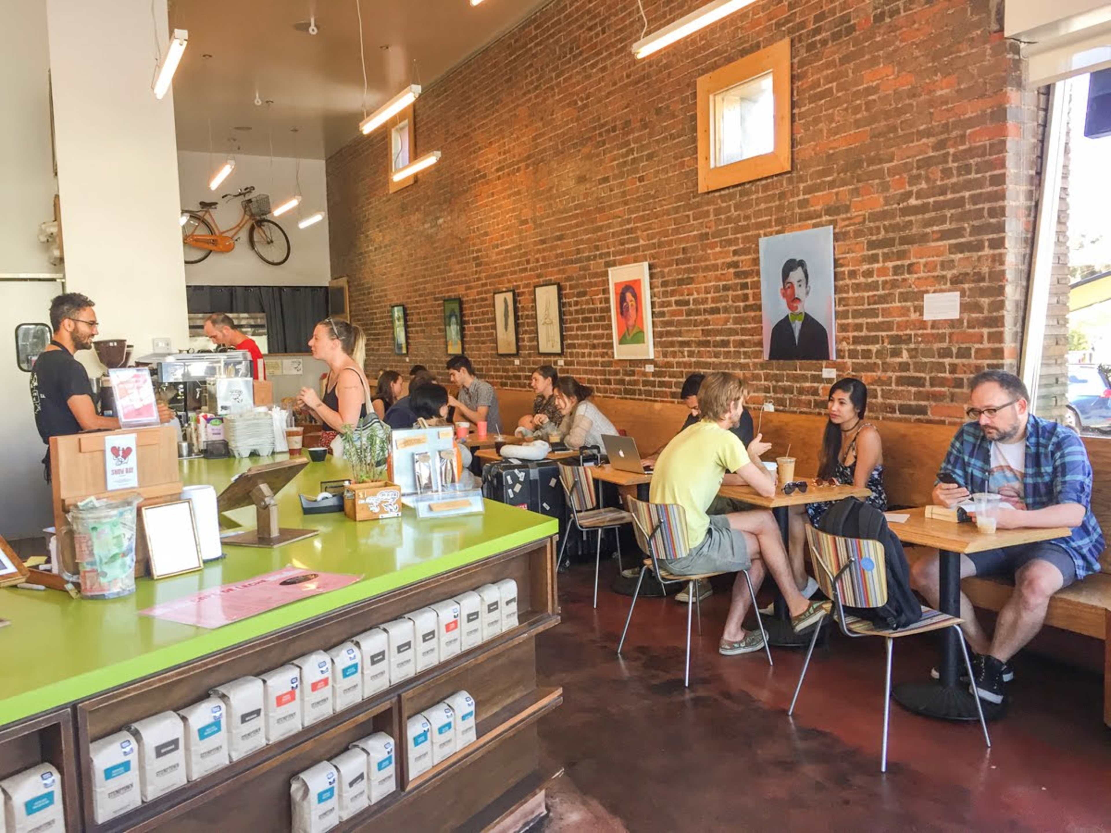A lively cafe with people sitting at tables, a service counter in the foreground, and artwork displayed on the exposed brick walls.