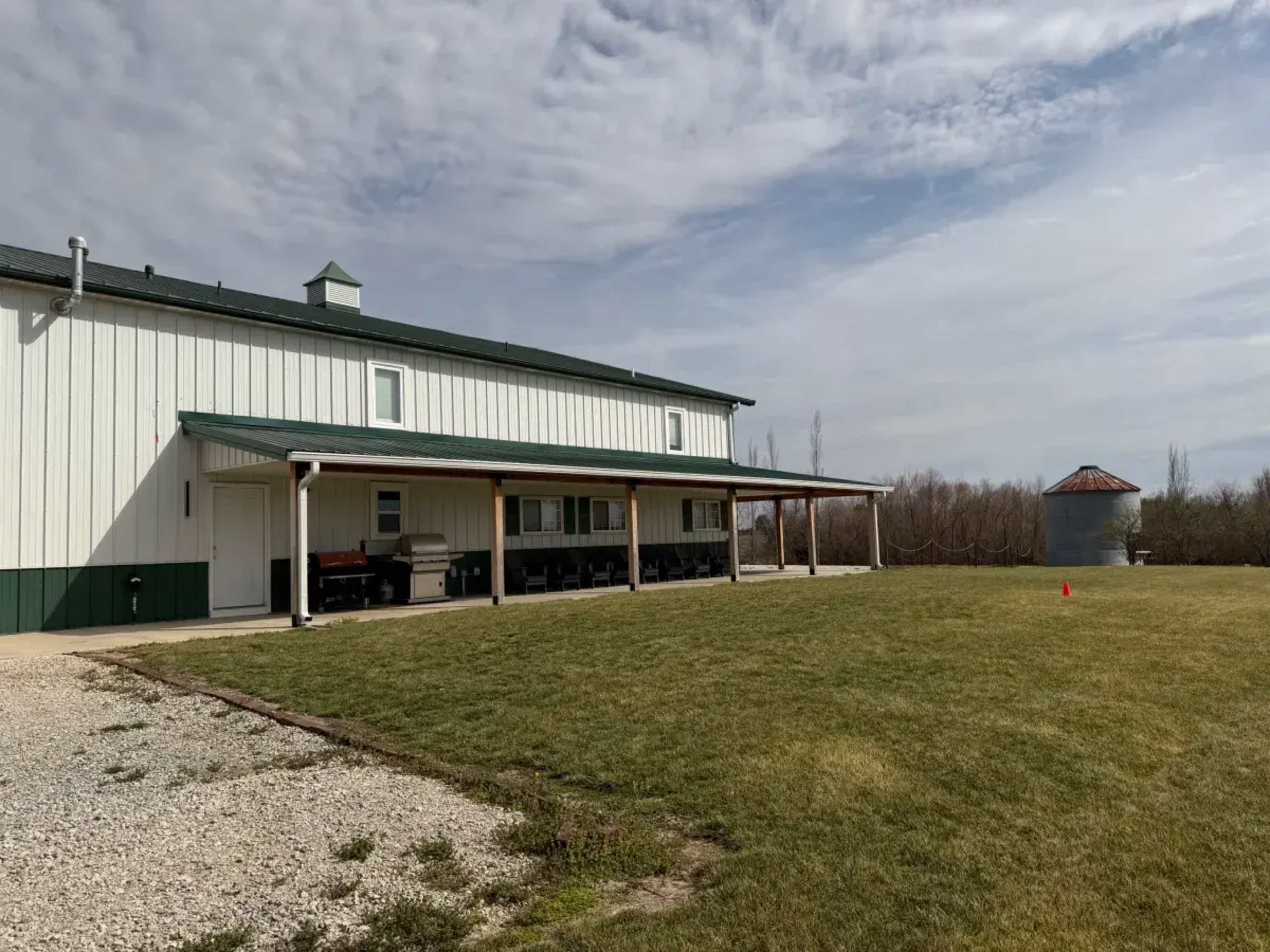 A large white building with green trim has a covered porch and is situated next to an open field, with a silos in the background.