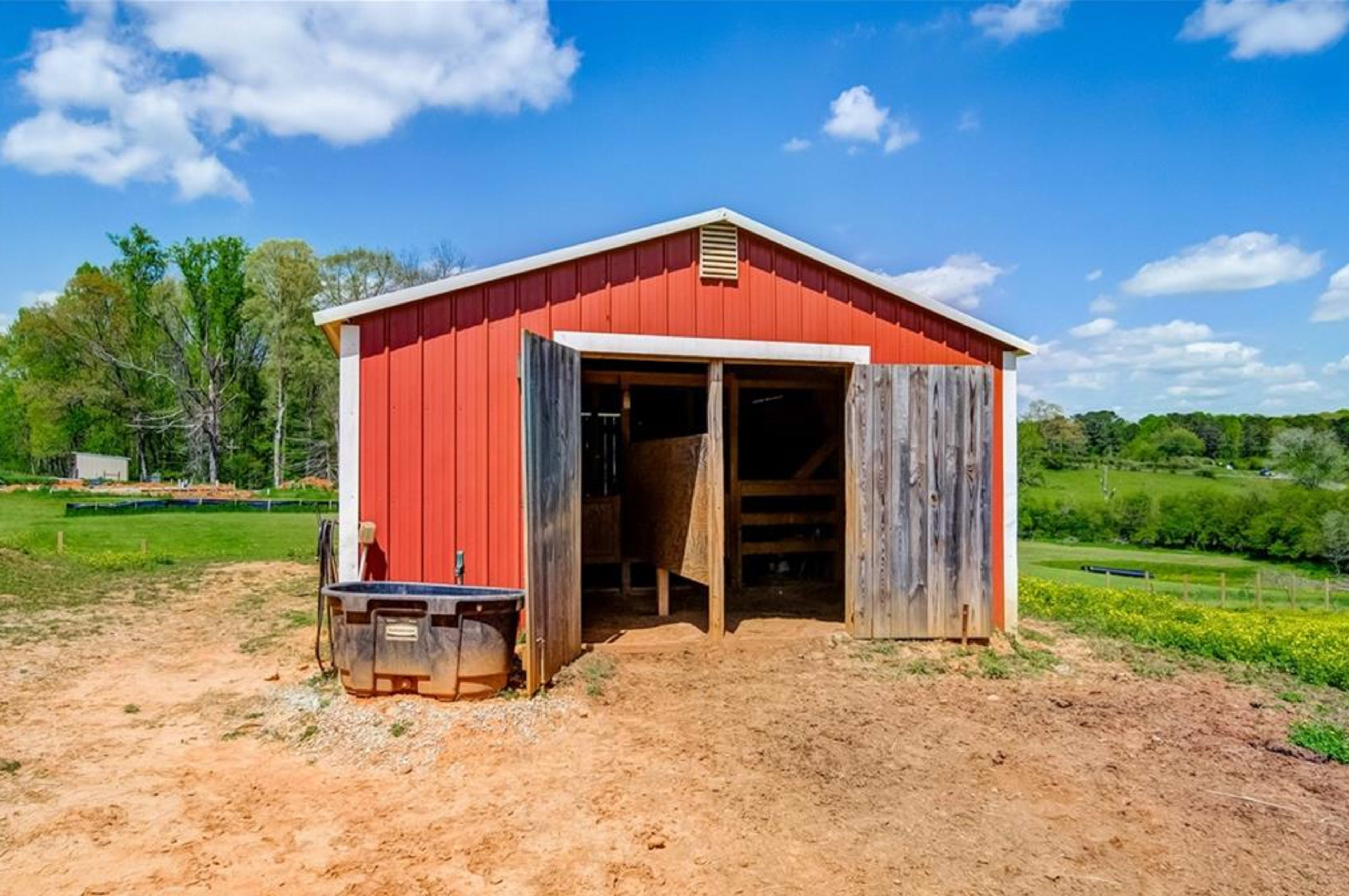 The image shows a red barn with open doors, situated in a rural landscape under a blue sky with scattered clouds.