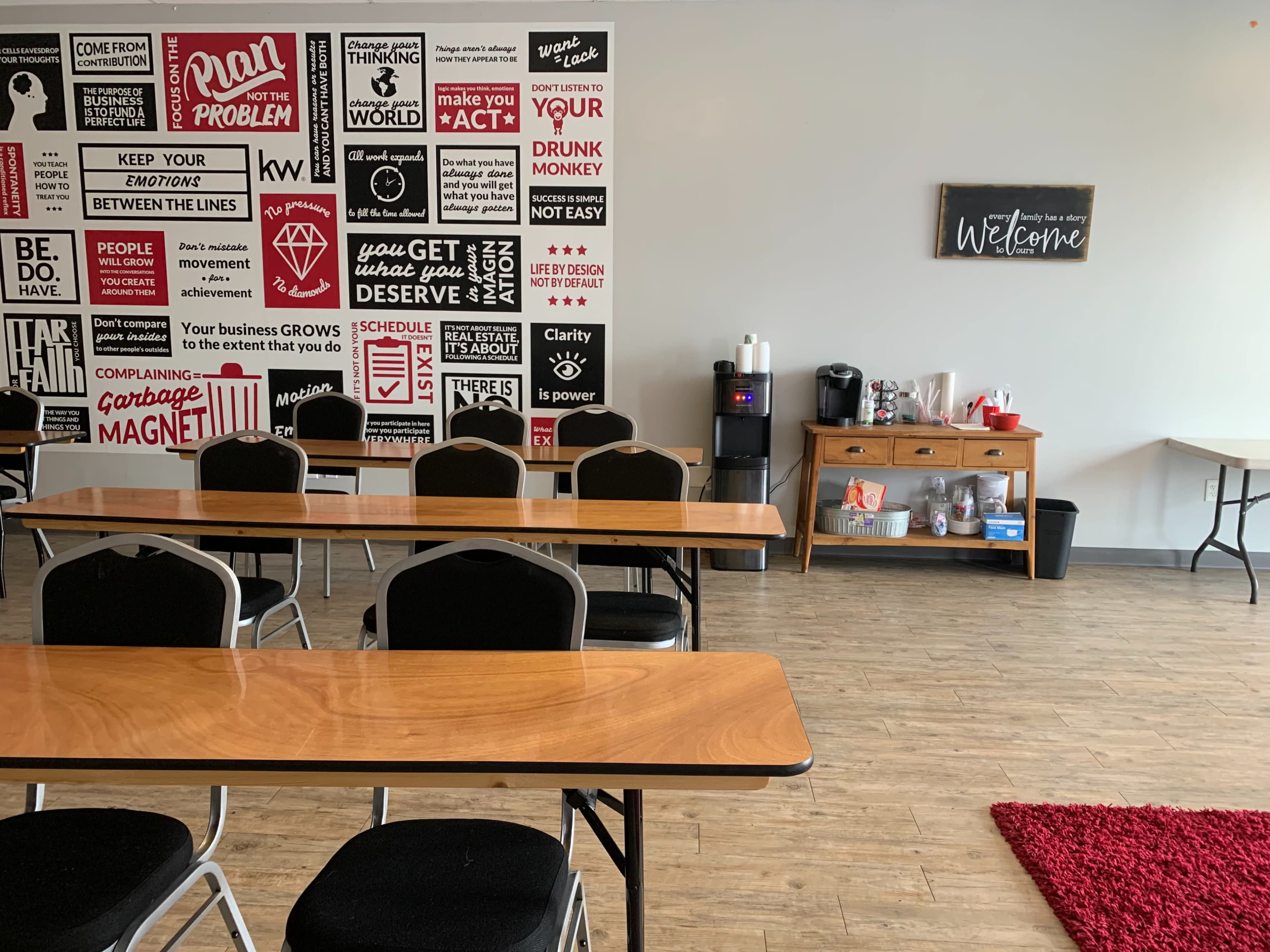 A spacious meeting room furnished with rows of tables and chairs, a water cooler, and a coffee station against a wall decorated with various motivational posters.