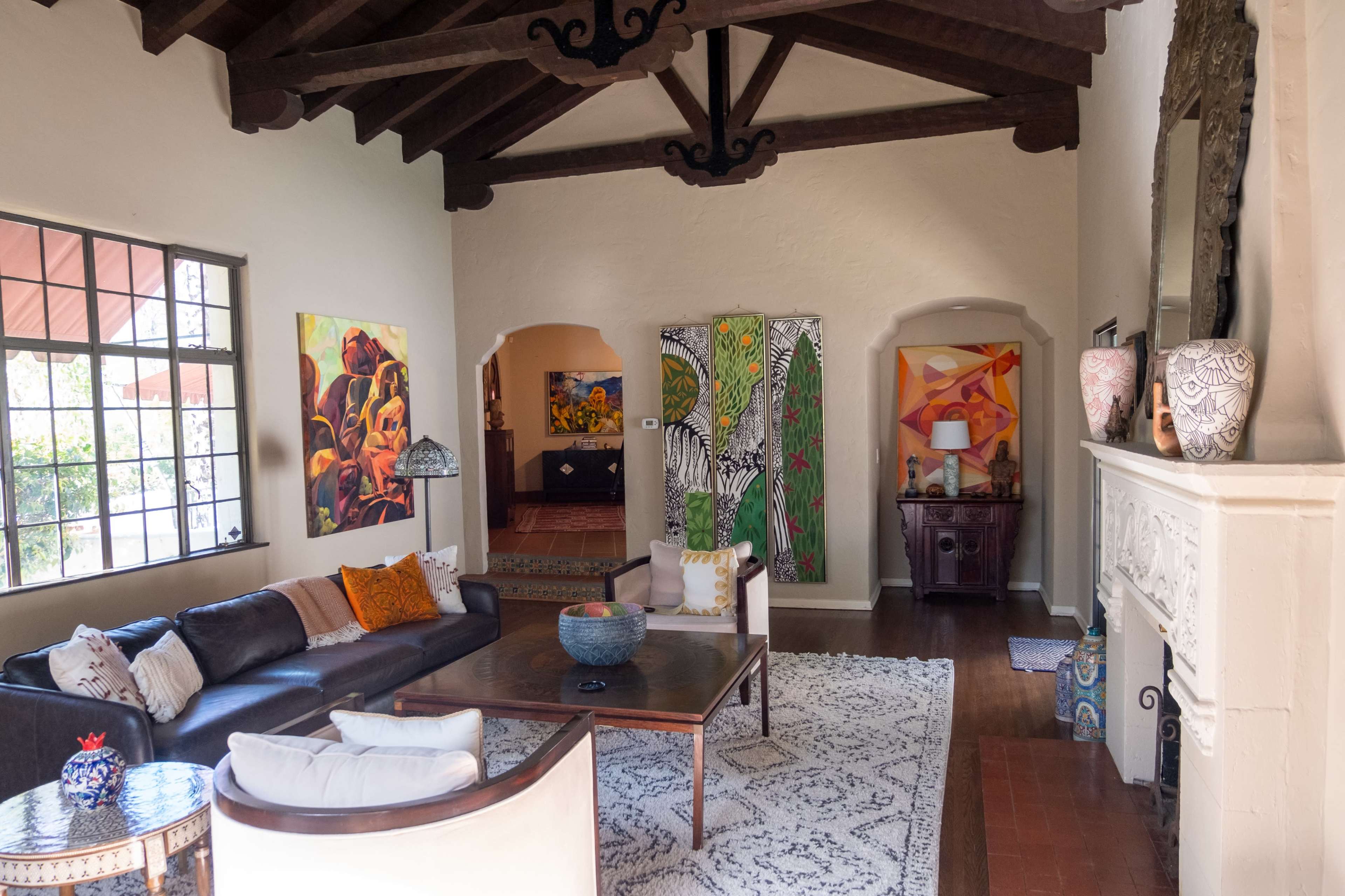 The image depicts a spacious living room with high wooden ceilings, two large paintings on the walls, a sectional sofa, a wooden coffee table, and a decorative fireplace.