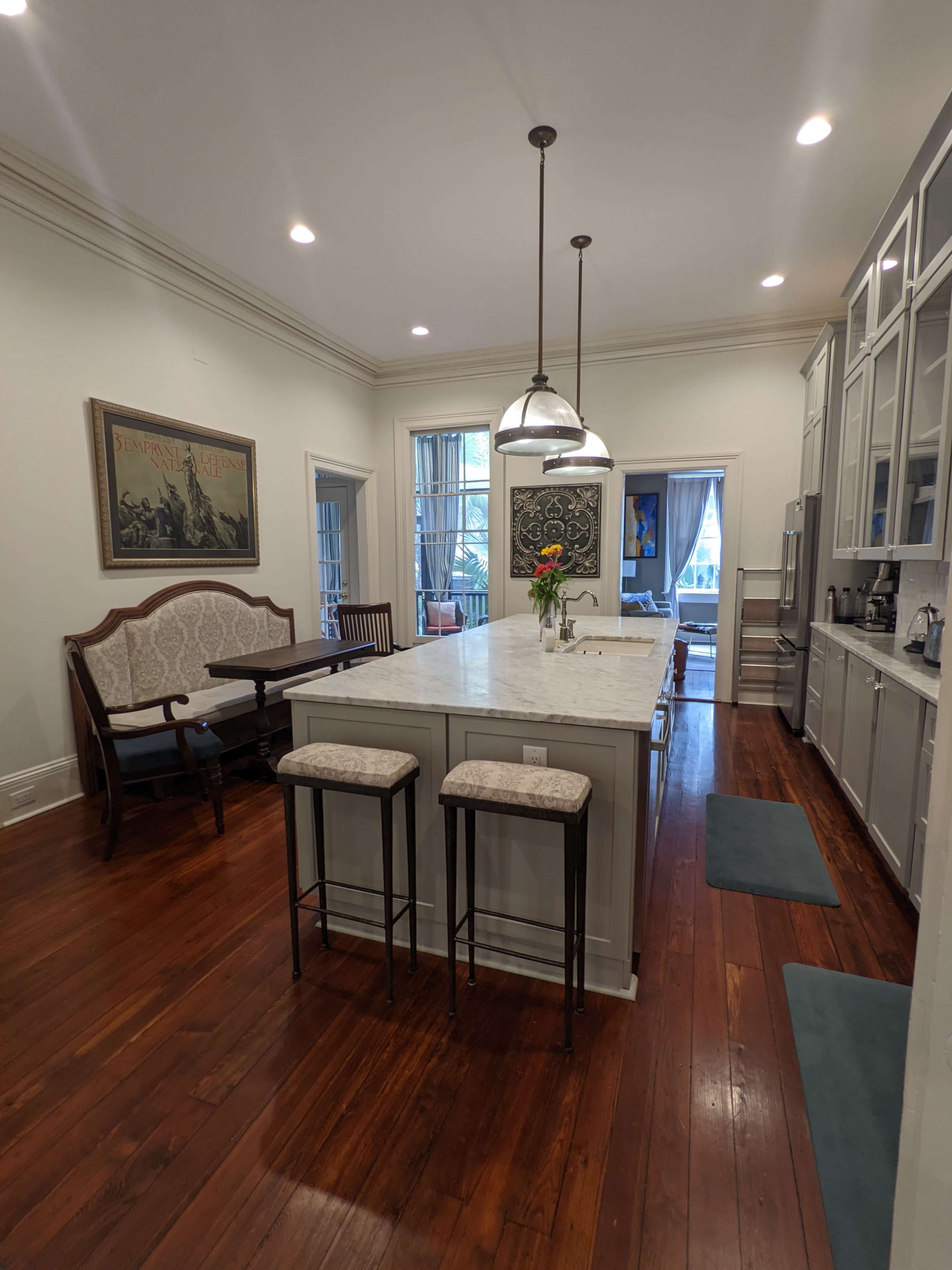 A modern kitchen with a central island, pendant lighting, and wooden floors, featuring a dining area with seating along one side.