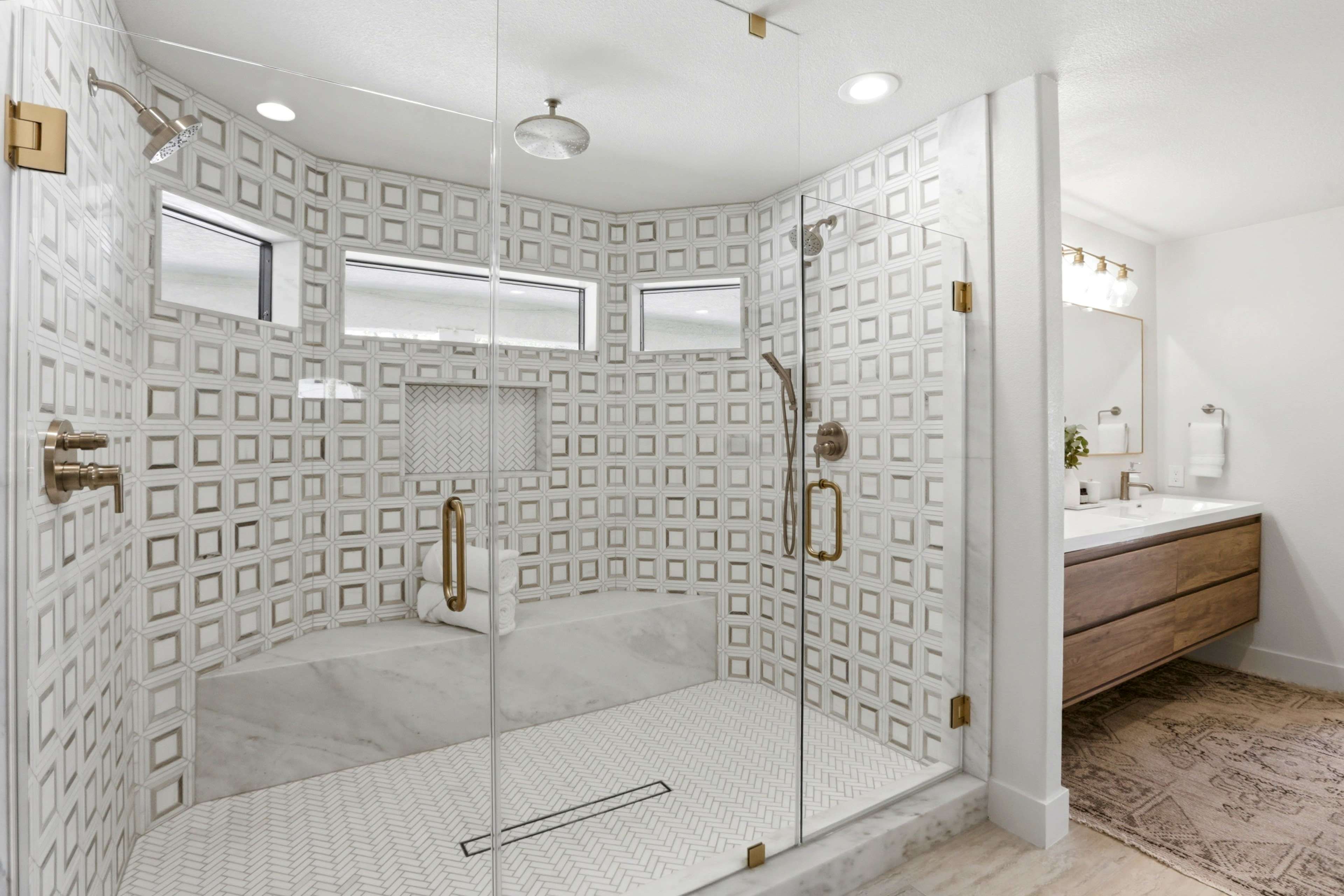 The image shows a modern bathroom featuring a walk-in shower with glass walls, intricate tile designs, and a wooden vanity with a sink.
