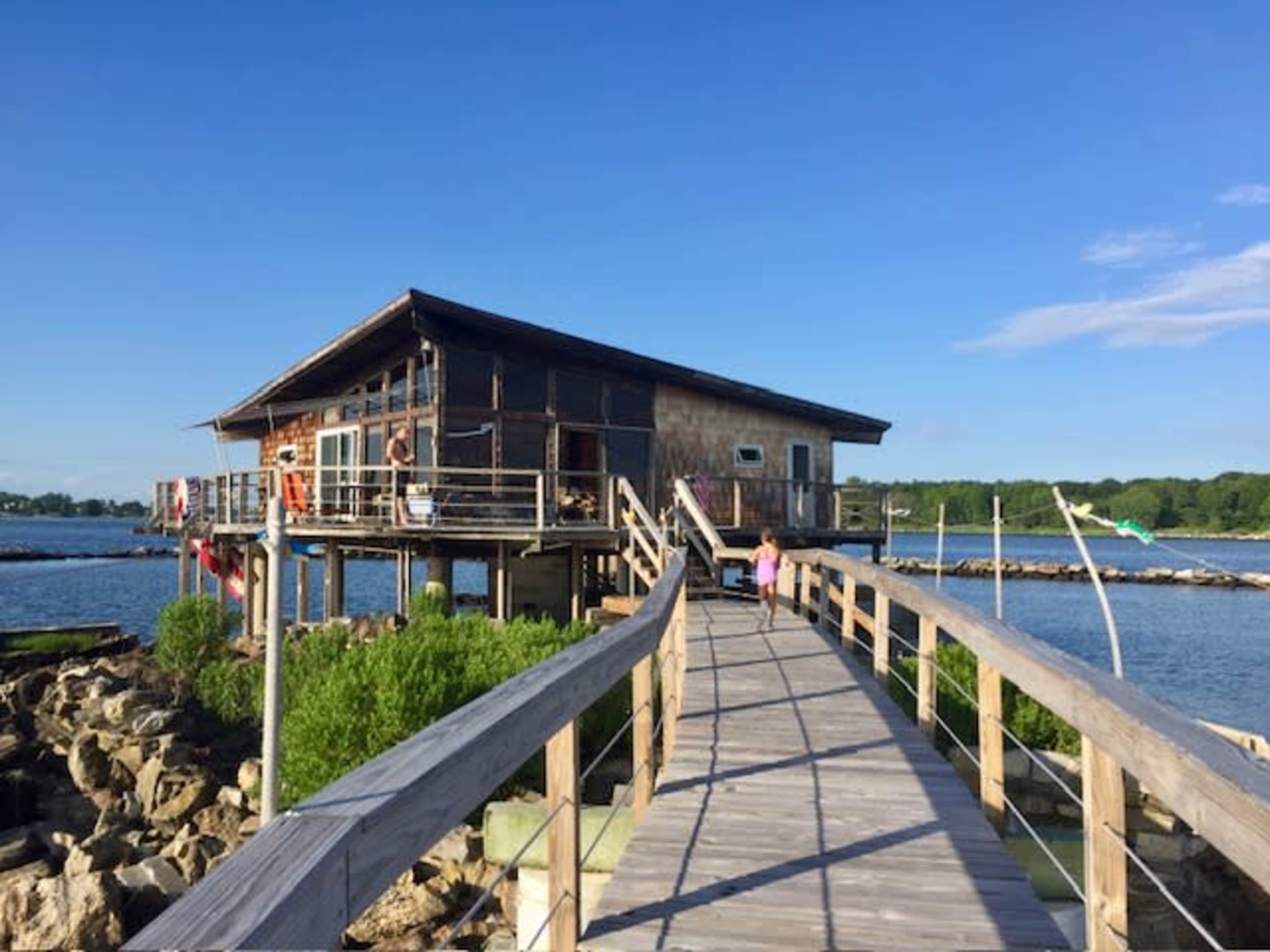 A wooden boardwalk leads to a lakeside cabin with large windows and a deck, surrounded by water and greenery.