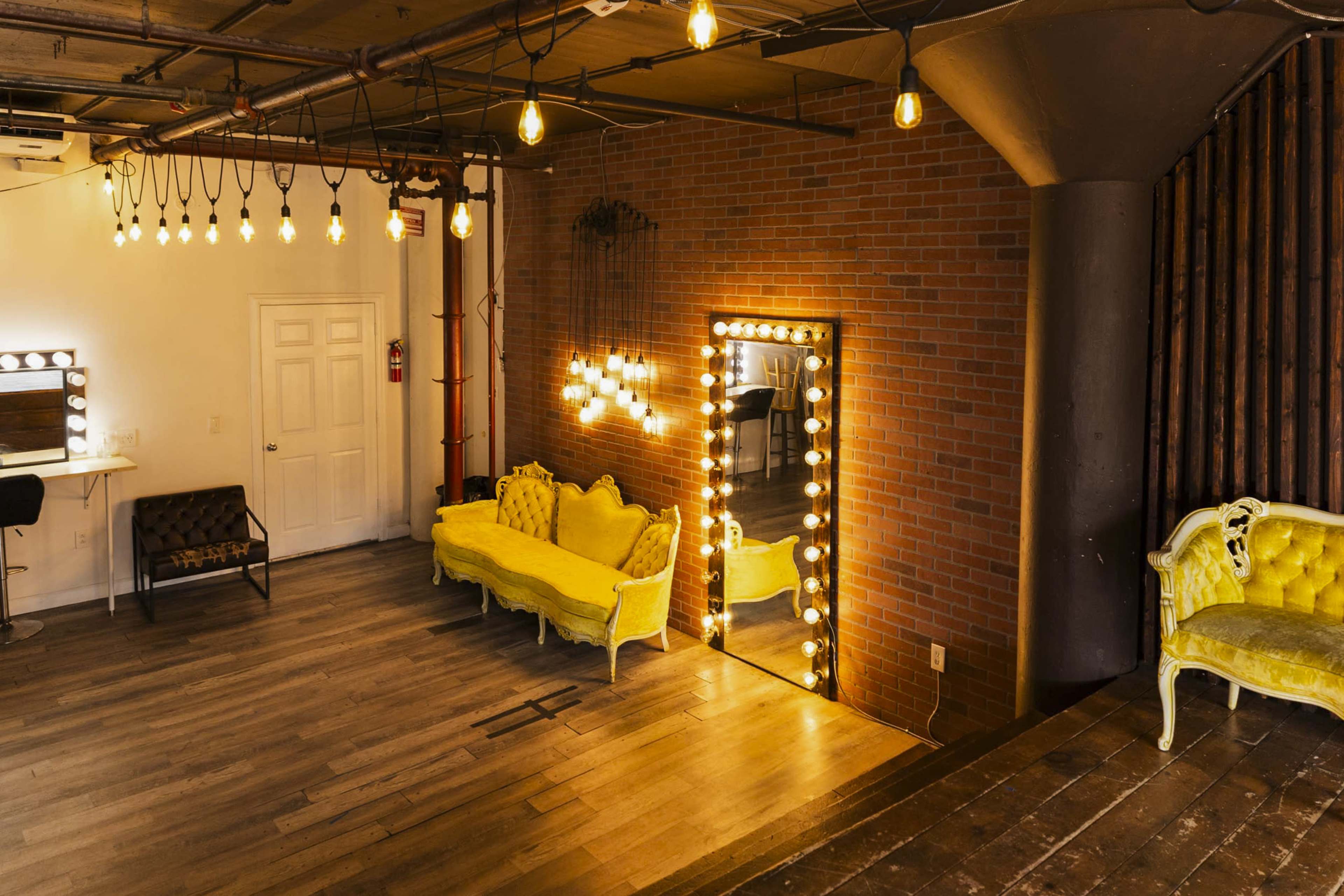 A stylish room with yellow sofas, a large mirror framed by lights, and exposed brick walls.