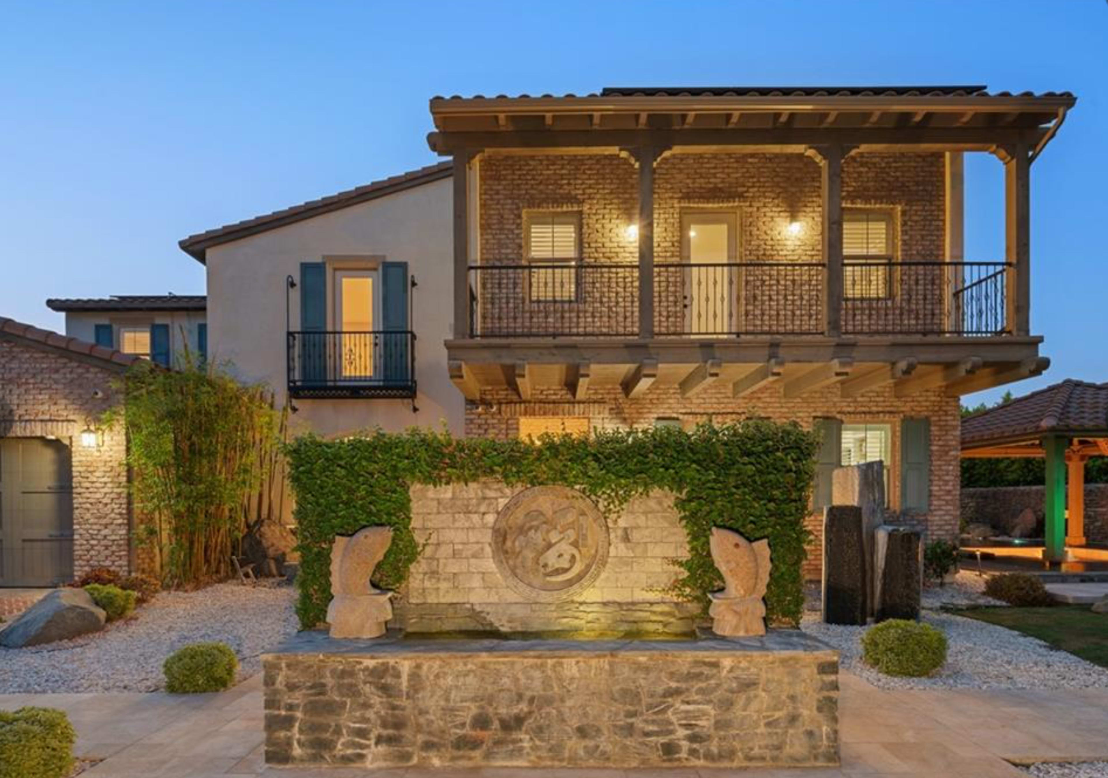 The image shows a two-story brick house with a stone fountain featuring sculptures in the foreground, surrounded by landscaped greenery and gravel pathways.