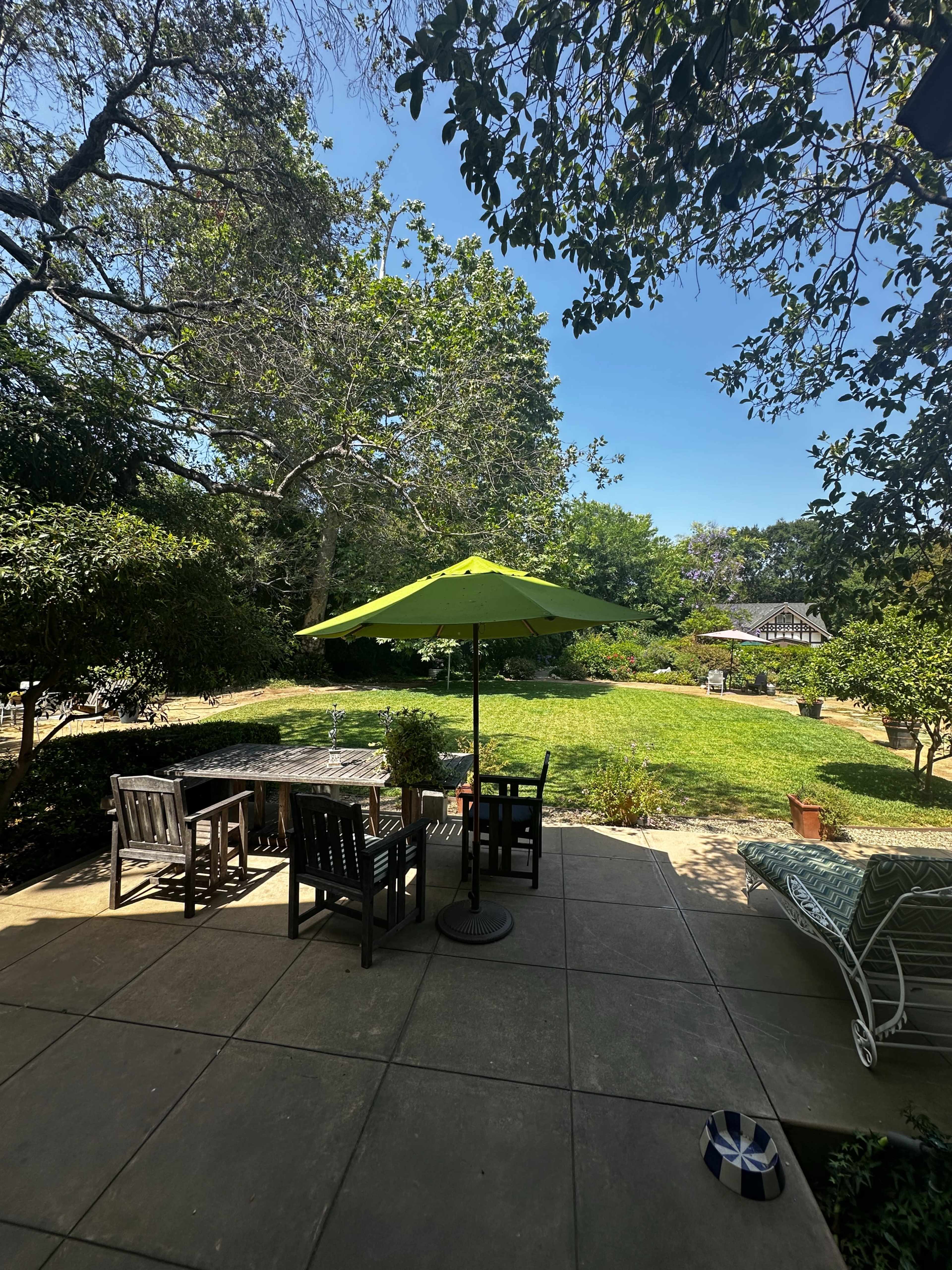 The image shows a patio area with outdoor chairs and a green umbrella, overlooking a grassy garden surrounded by trees.