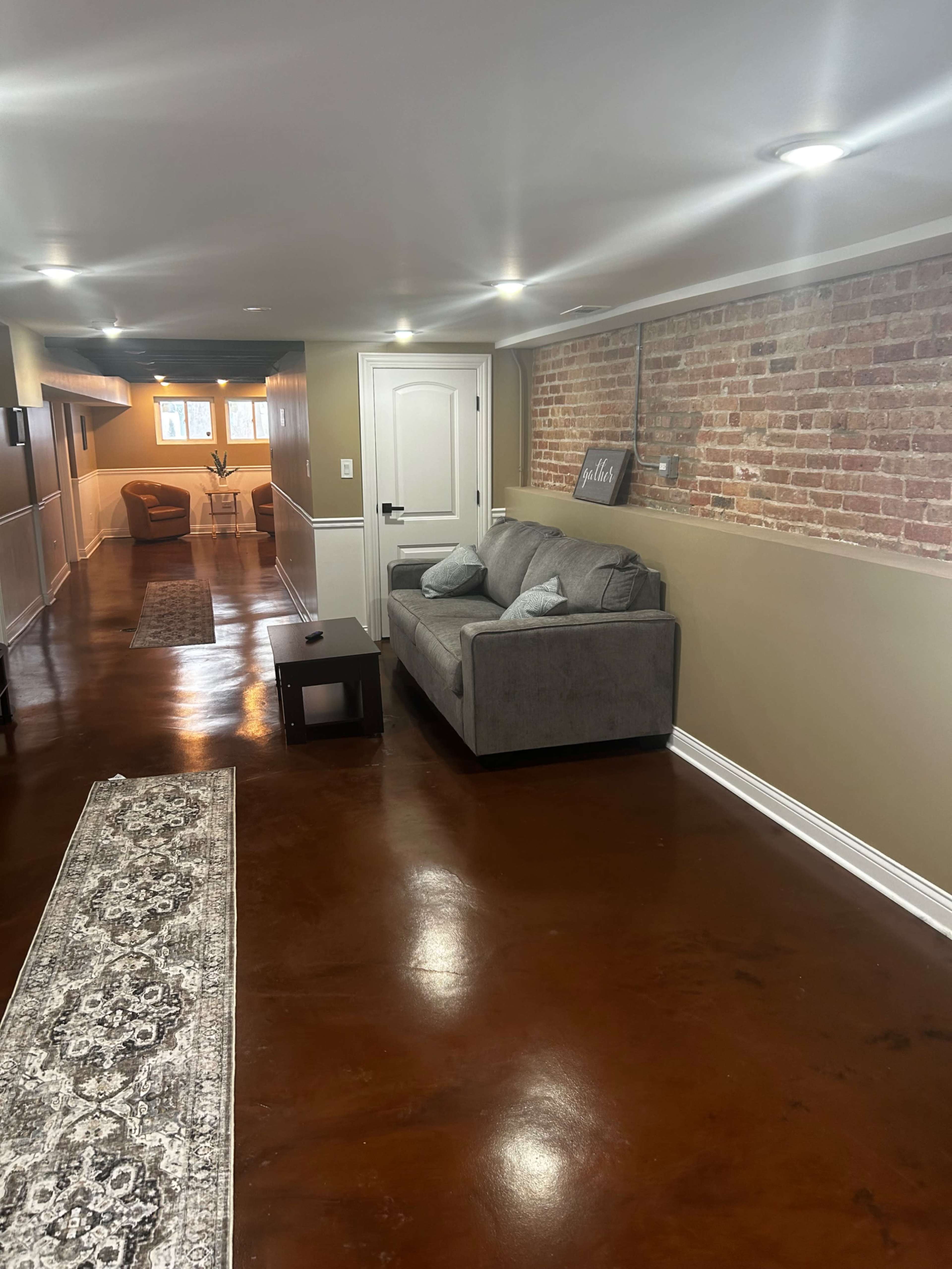 The image shows a hallway with a gray couch and a small table, featuring a brick wall and polished concrete flooring.