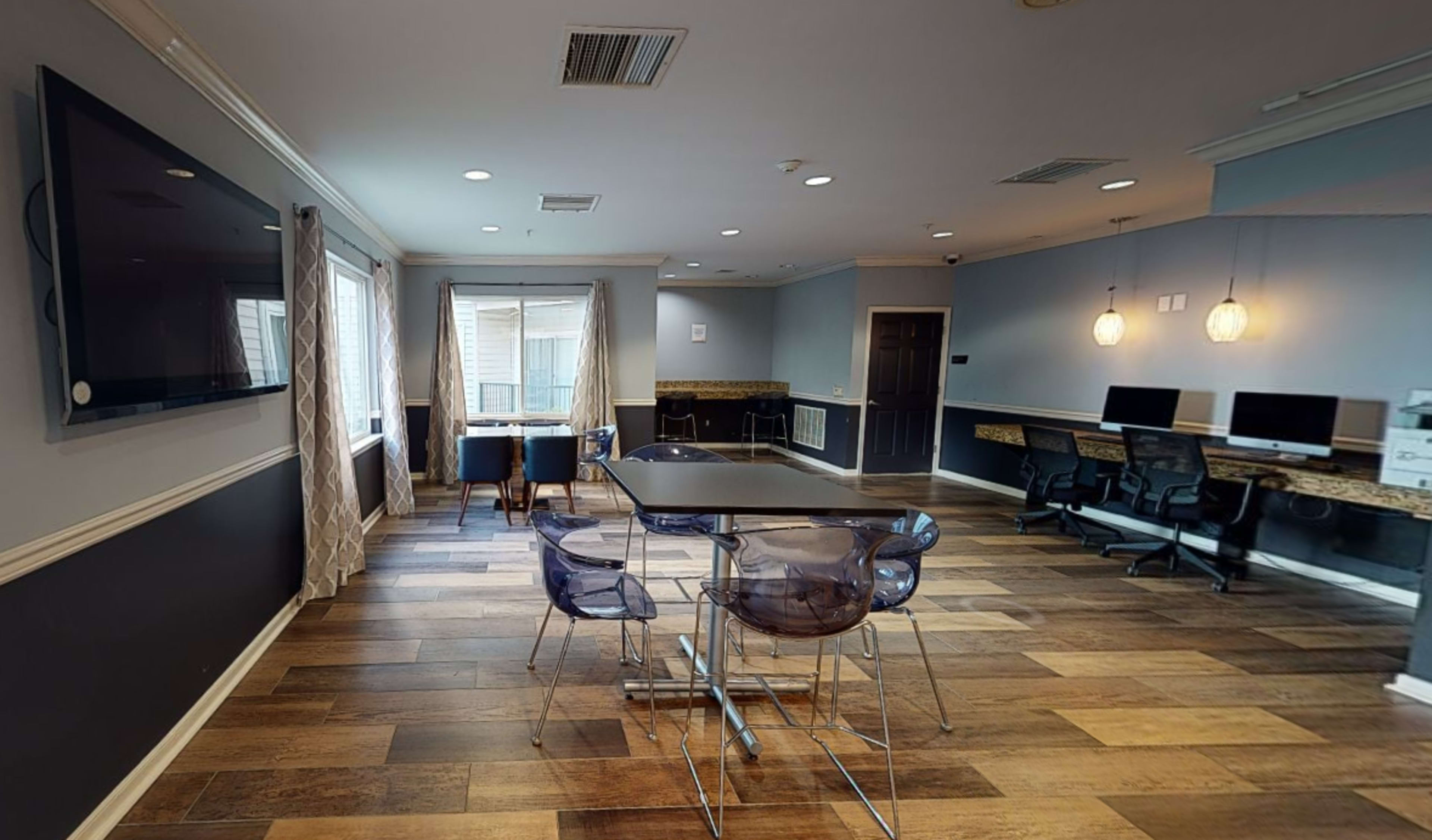 A modern room features a large table surrounded by transparent chairs, with a television on one wall and workstations in the background.