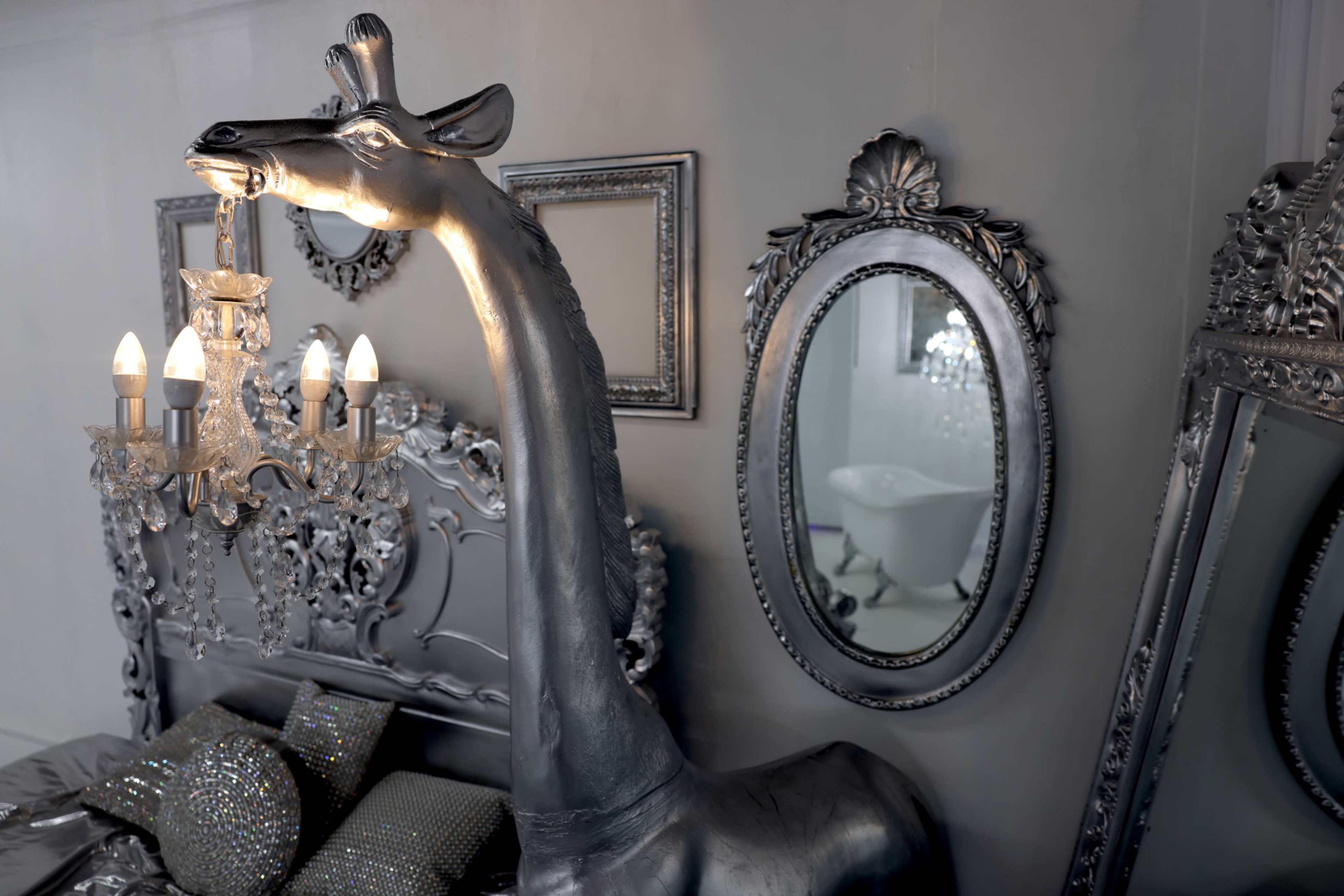 The image features a silver giraffe sculpture beside an ornate mirror and a chandelier in a stylish interior setting.