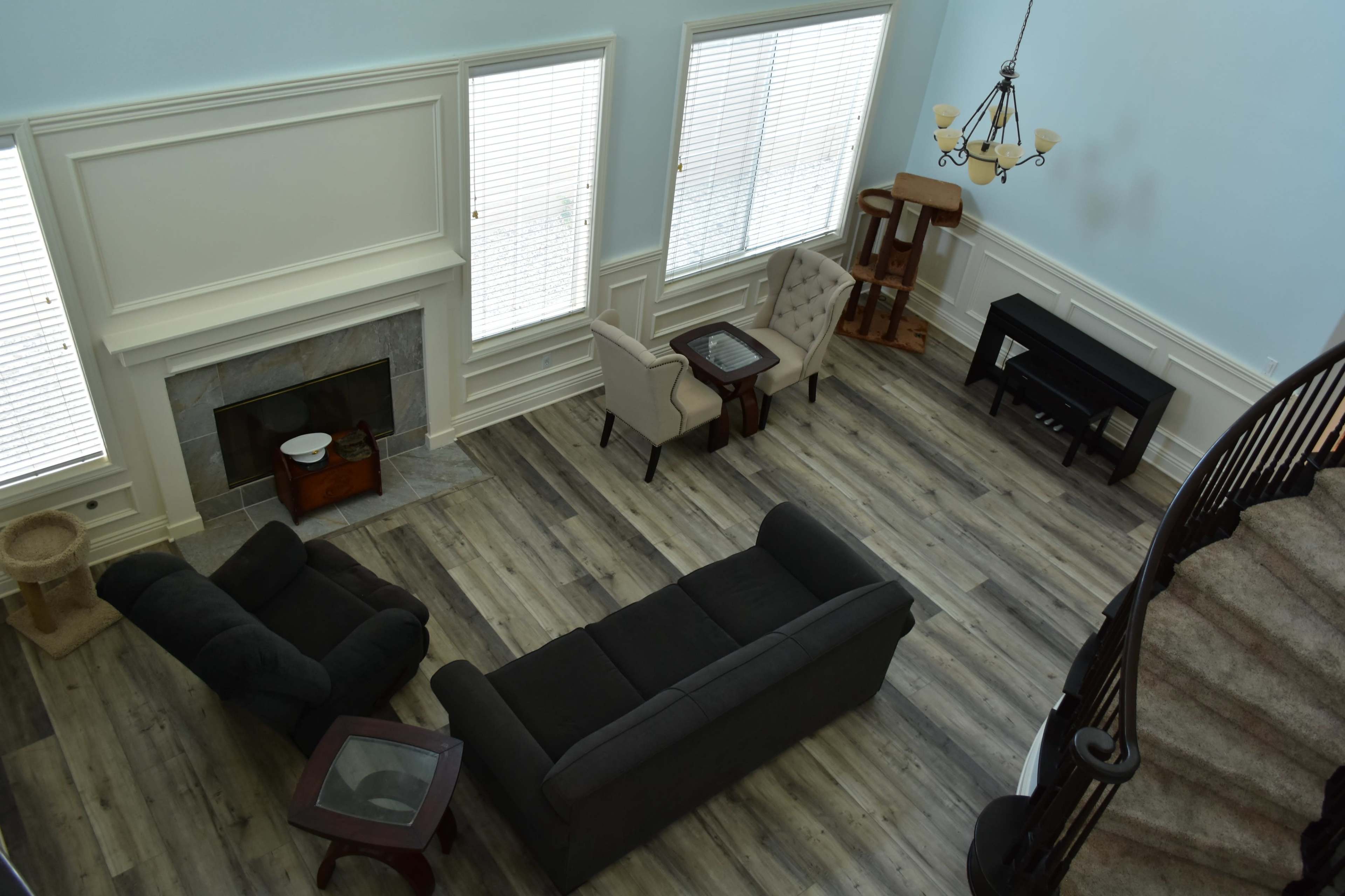 A spacious living room with a gray sofa, two armchairs, a coffee table, a side table, and a staircase leading to an upper level, all set against light blue walls and large windows.