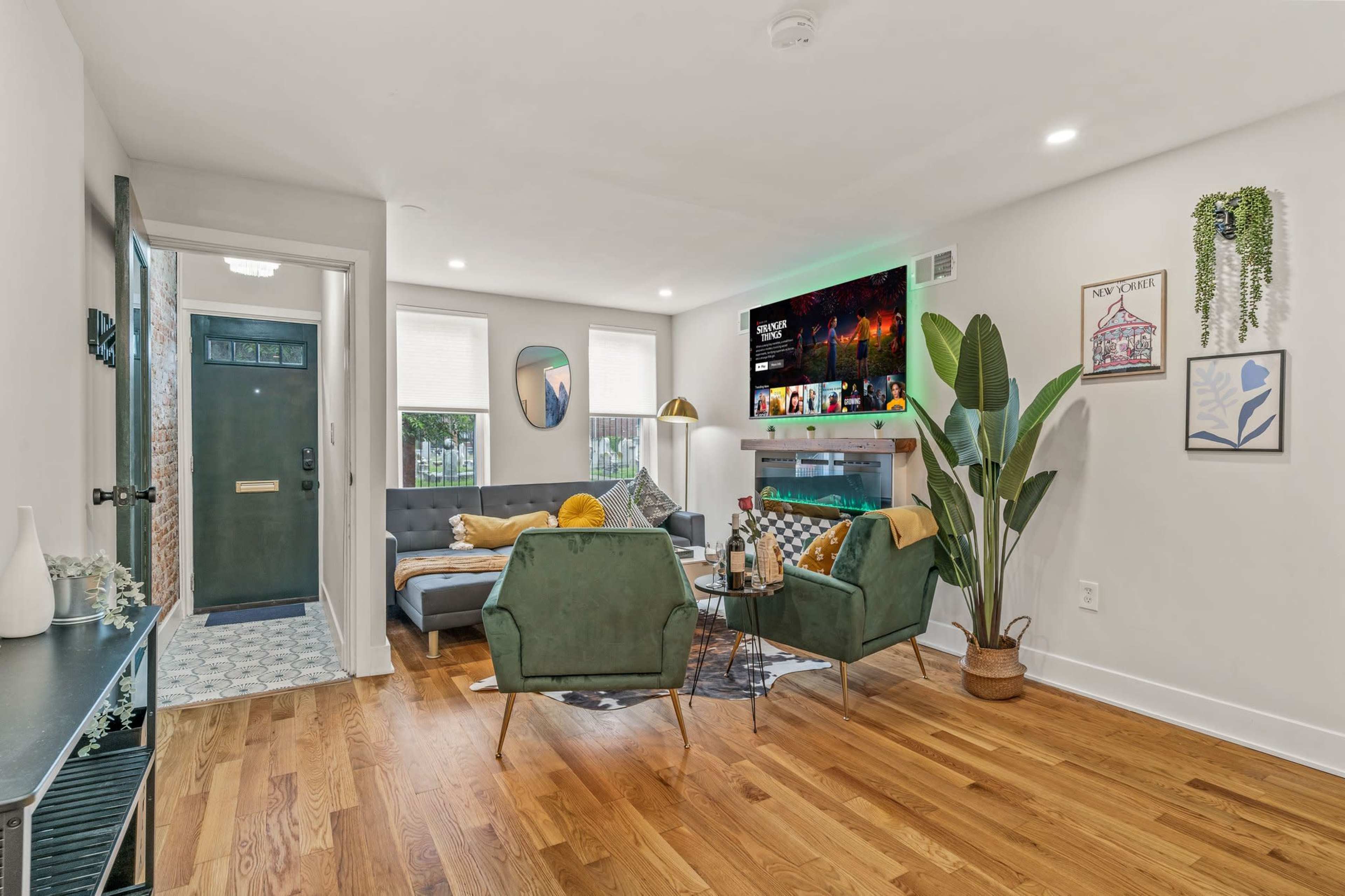 The living room features a gray sofa, two green armchairs, a modern entertainment unit with a television, and a large plant in a corner, all situated on a hardwood floor.