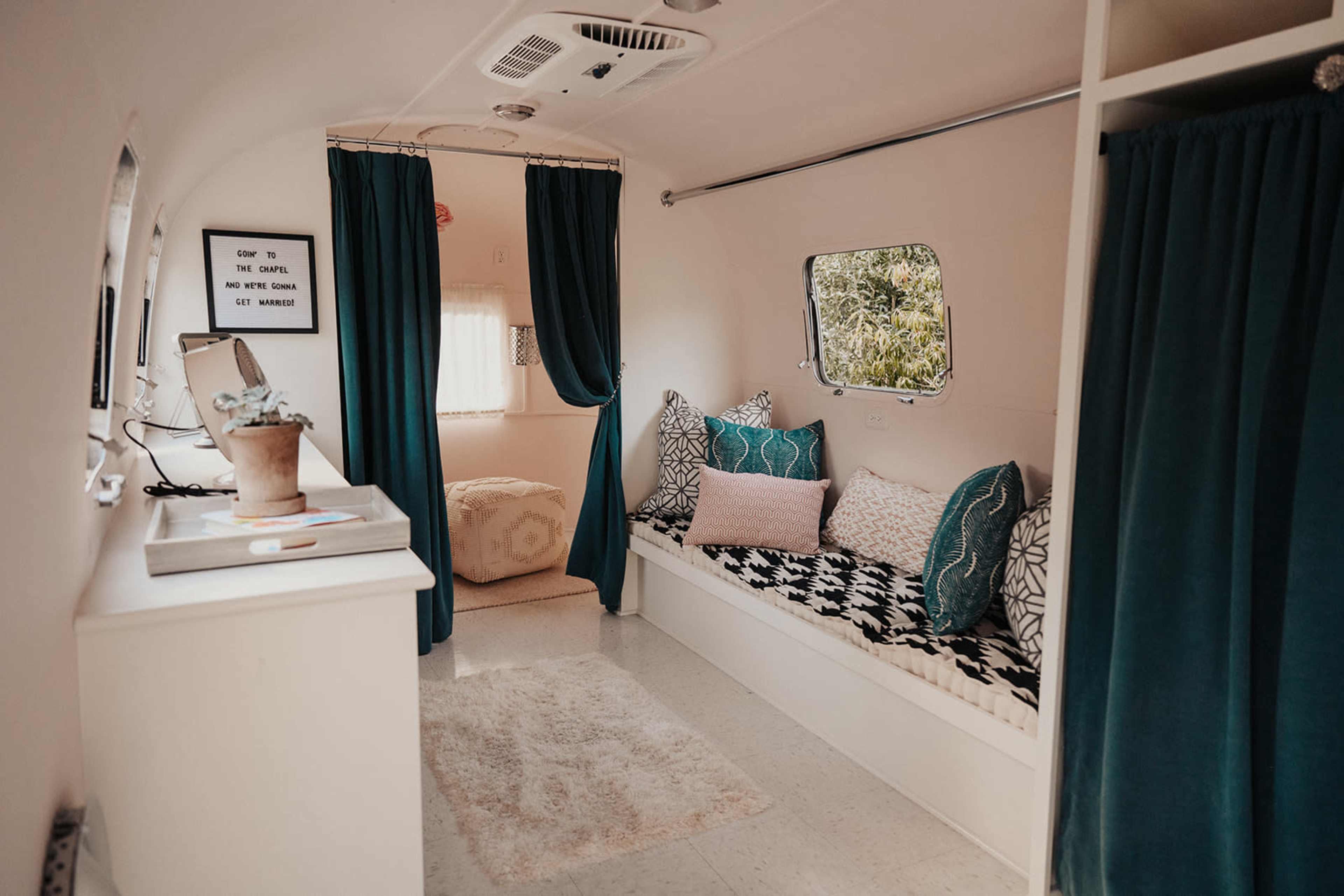 A small retro-style trailer interior featuring a cozy seating area with pillows, a window, and teal curtains.