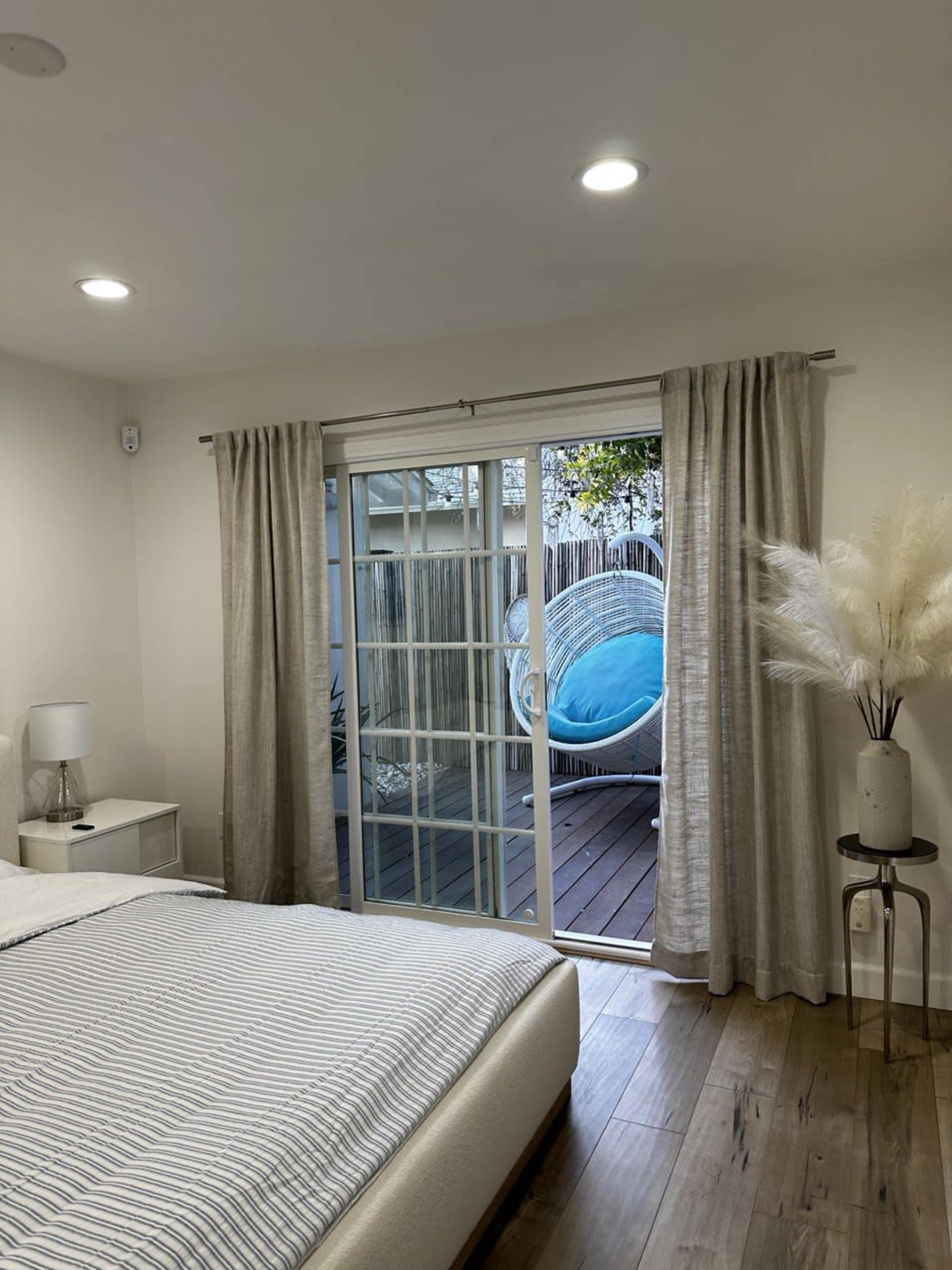 A well-lit bedroom features a bed with a striped blanket and a sliding glass door that opens to a deck with a hanging chair.