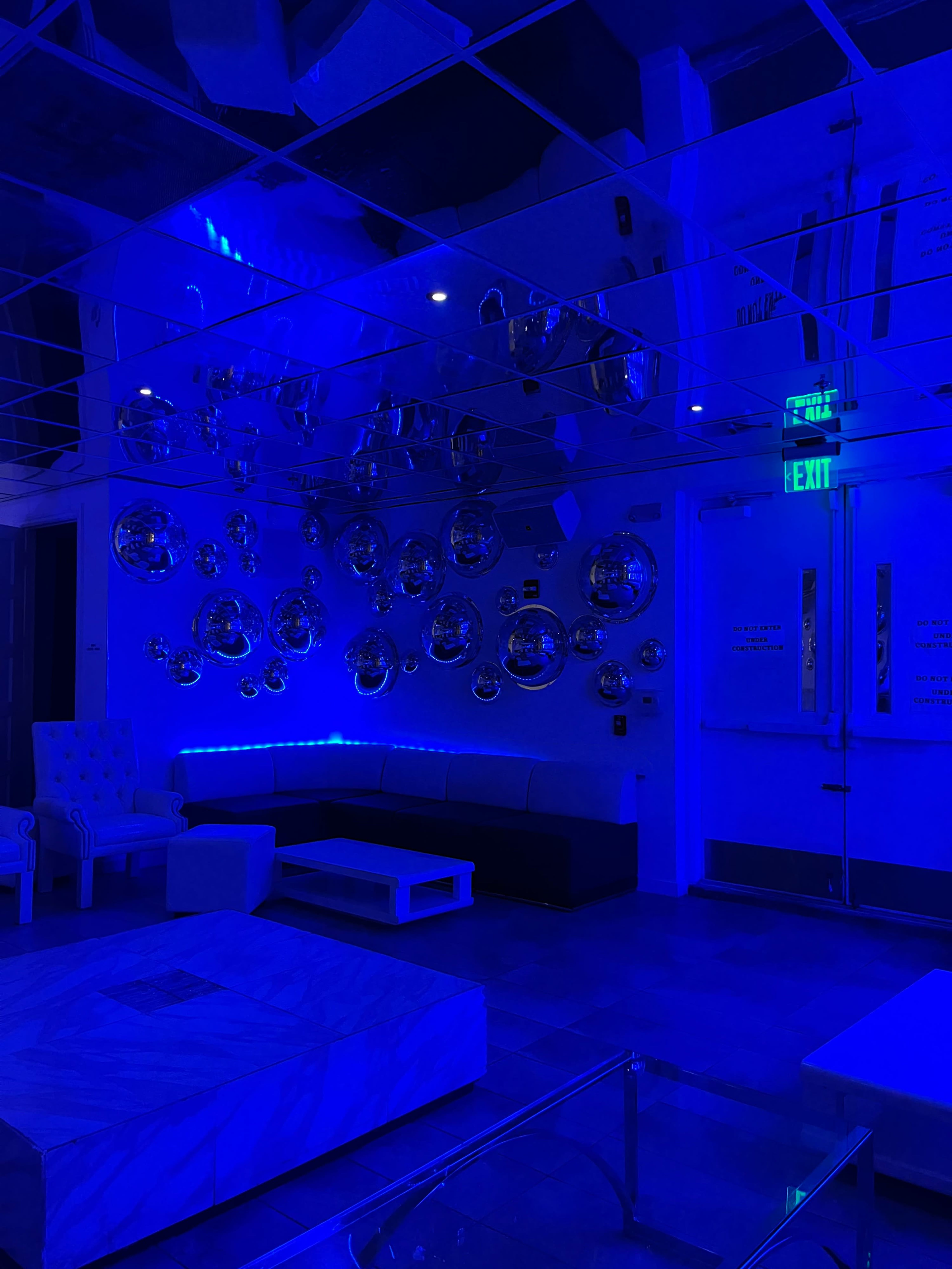 The image shows a modern lounge area with blue lighting, featuring a couch, a coffee table, and a wall adorned with reflective spherical decorations.