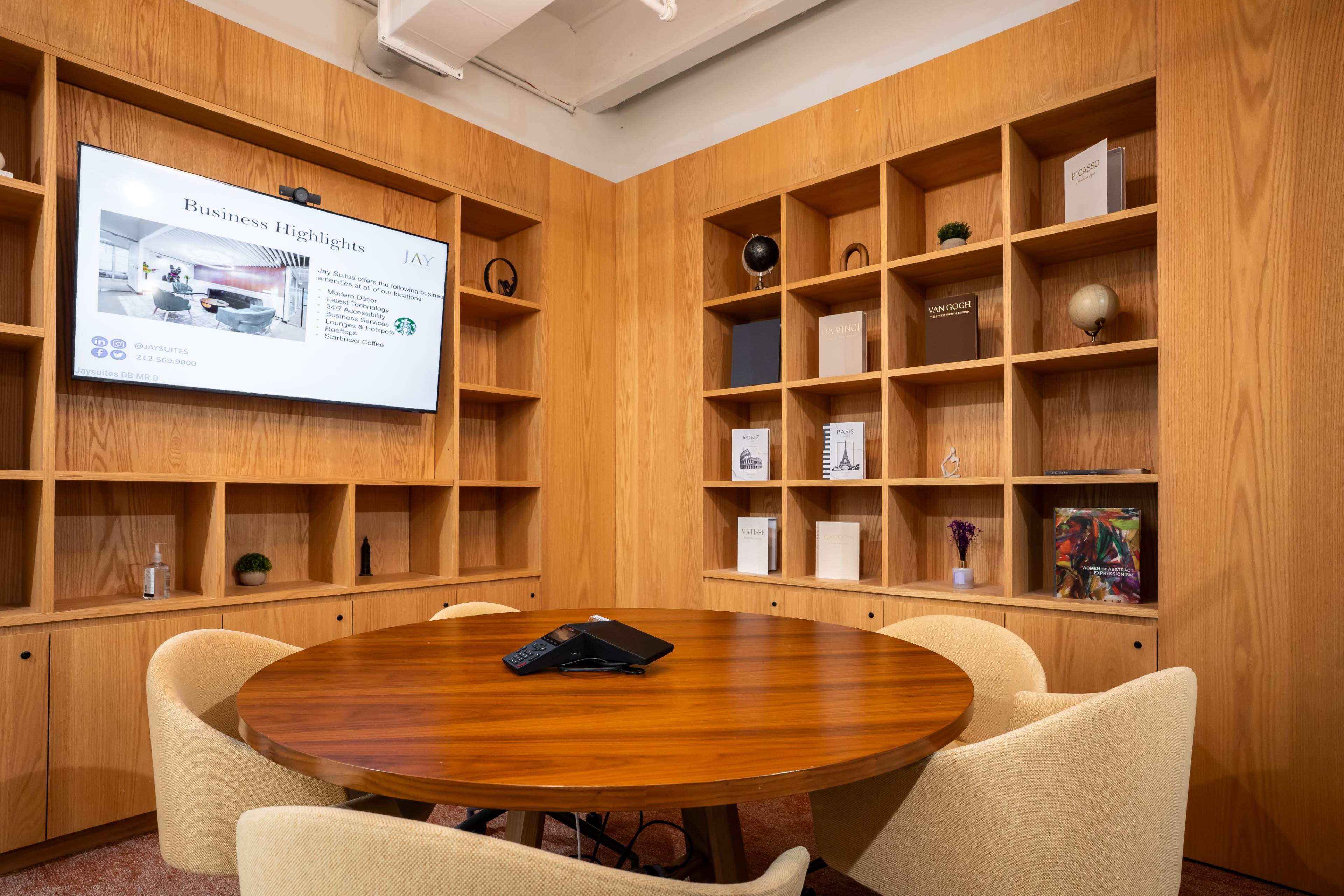 A conference room features a round wooden table surrounded by four upholstered chairs, with a large screen displaying business information on one wall and wooden shelves filled with books and decorative items on another.