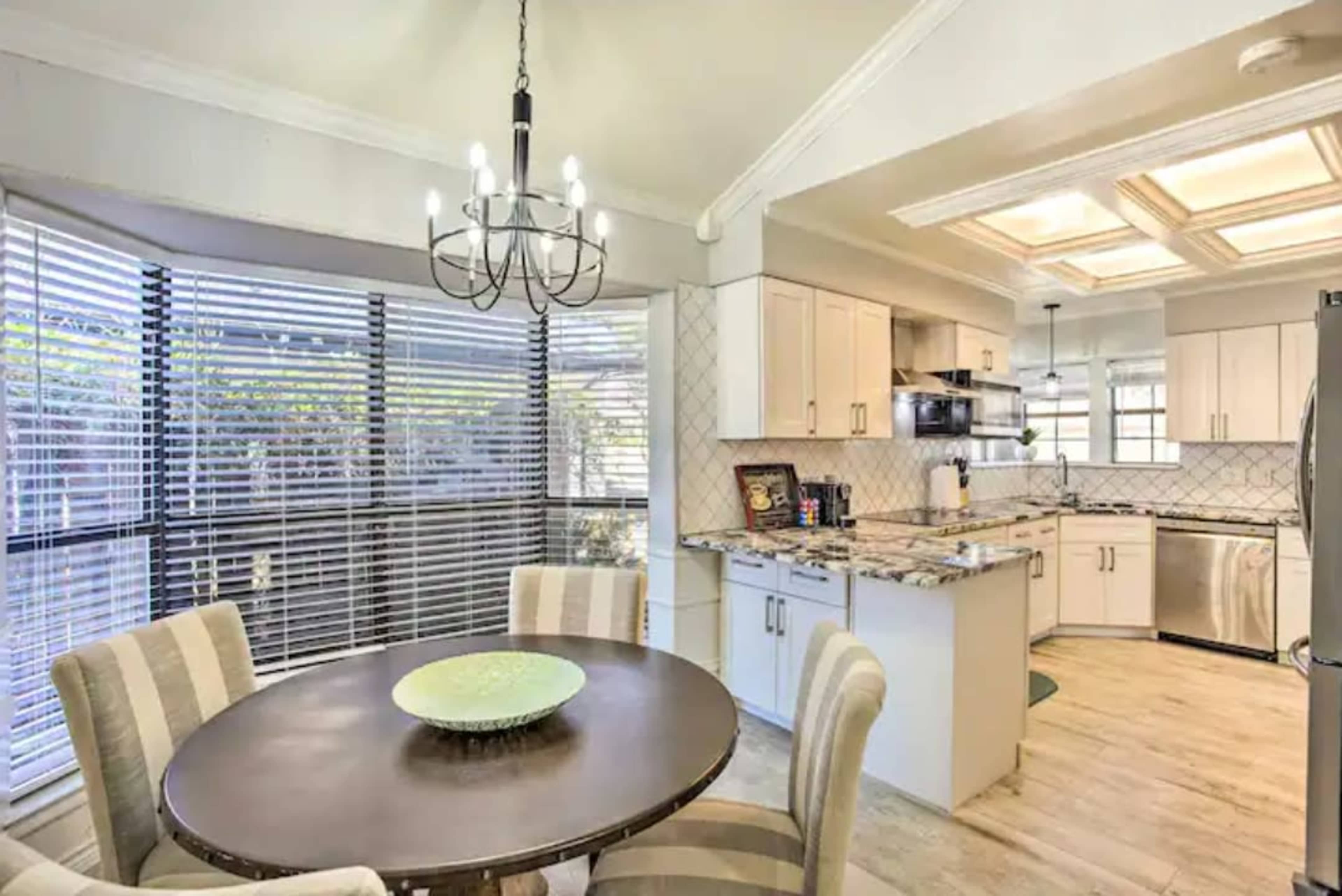 The image shows a modern kitchen and dining area with a round table, four chairs, and large windows covered with blinds.