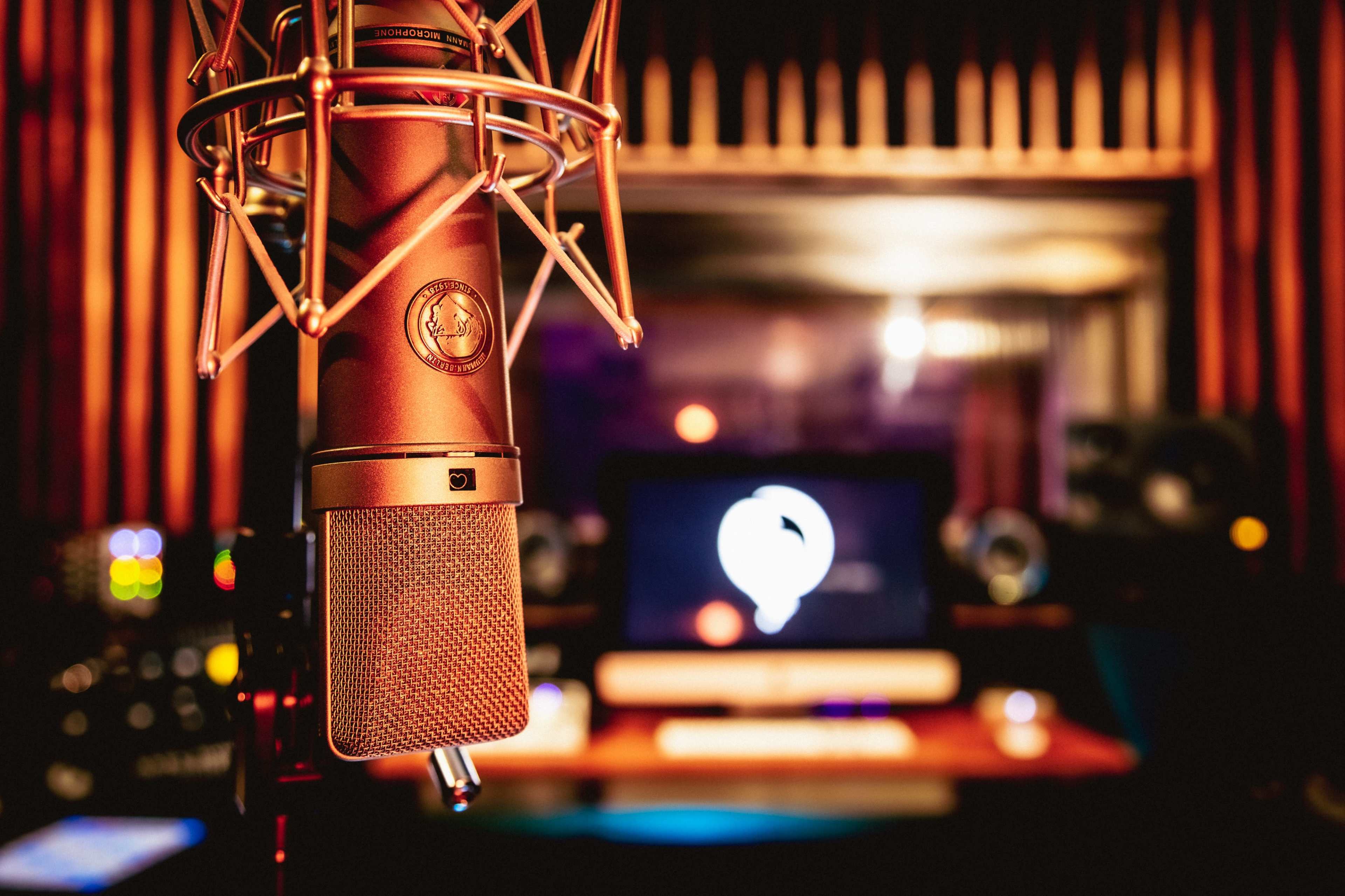 A microphone is positioned prominently in a sound studio with audio equipment and a computer in the background.
