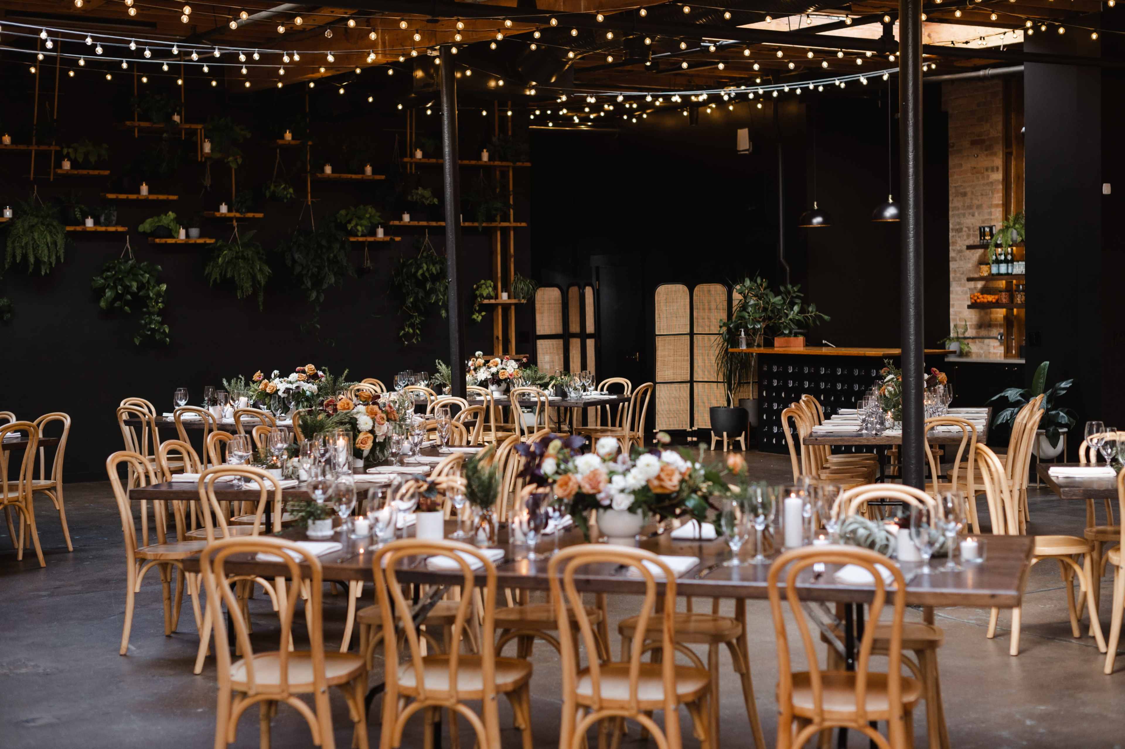 A dimly lit event space features long wooden tables elegantly arranged with floral centerpieces and surrounded by bentwood chairs, with string lights overhead and greenery adorning the walls.