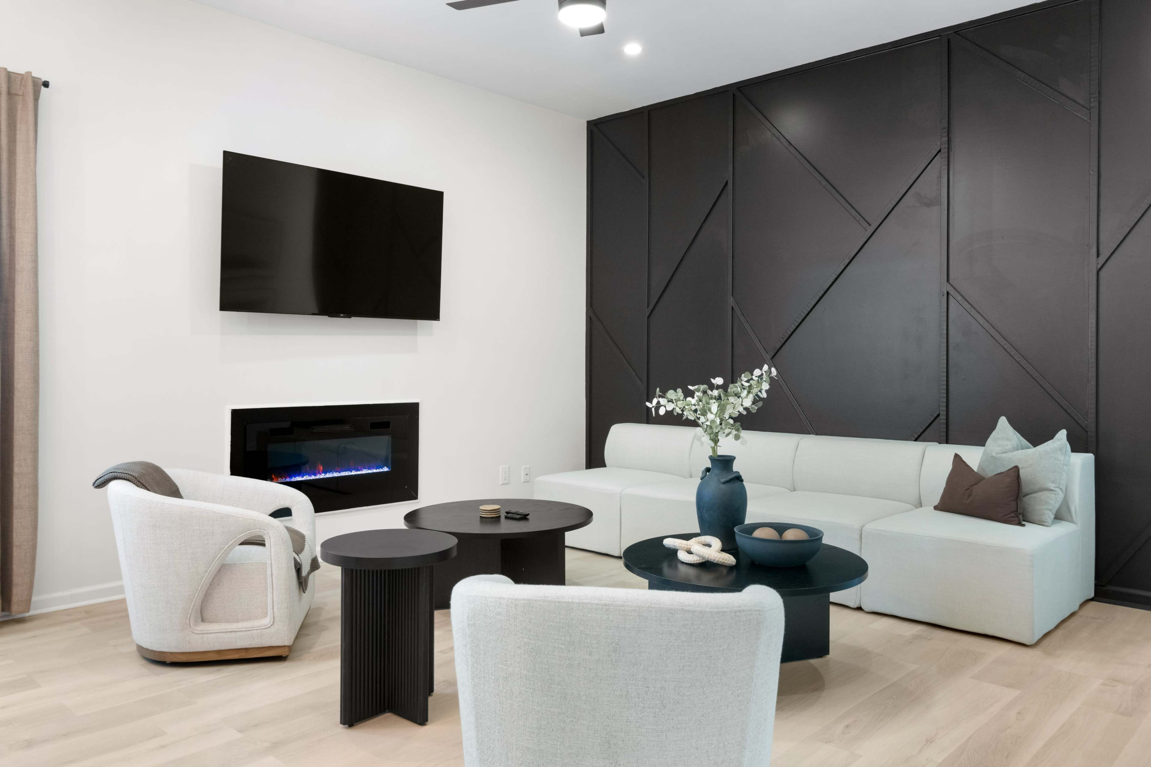 The image depicts a modern living room featuring a light-colored sectional sofa, a wall-mounted television, and a sleek electric fireplace, with two chairs and various decorative elements on the tables.