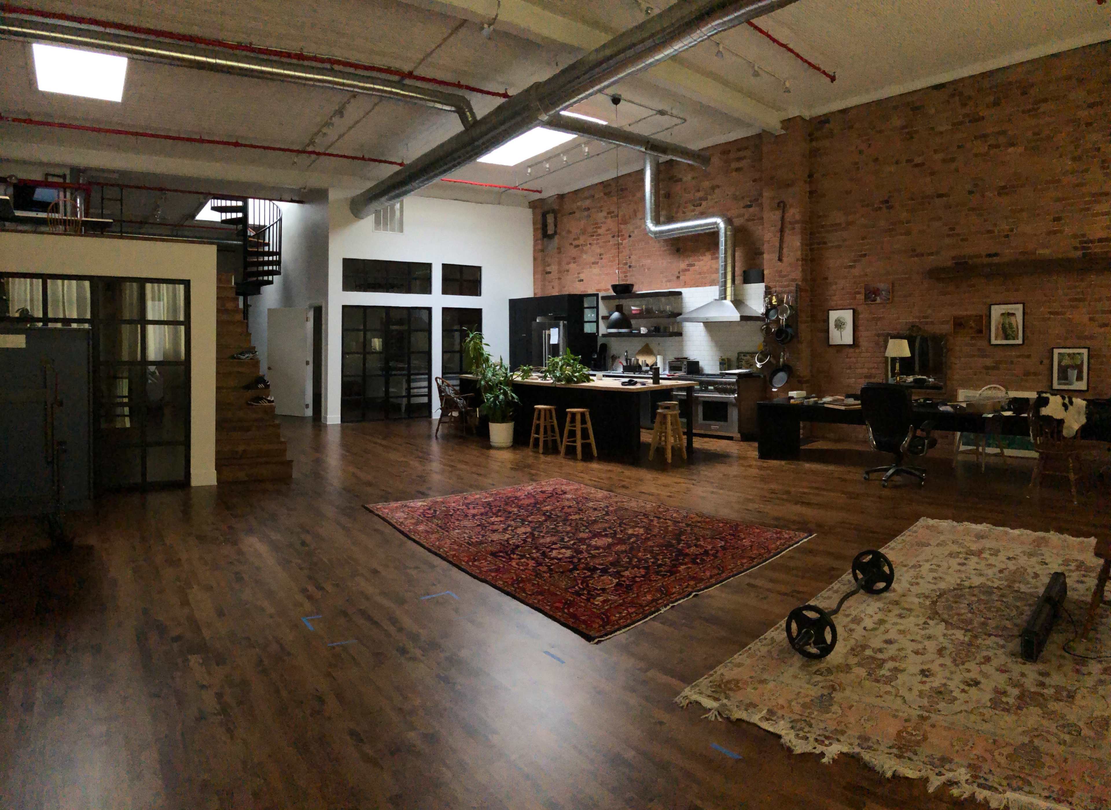 The image shows a spacious loft-style interior with exposed brick walls, a kitchen area, a dining space, and a living room featuring rugs and workout equipment.