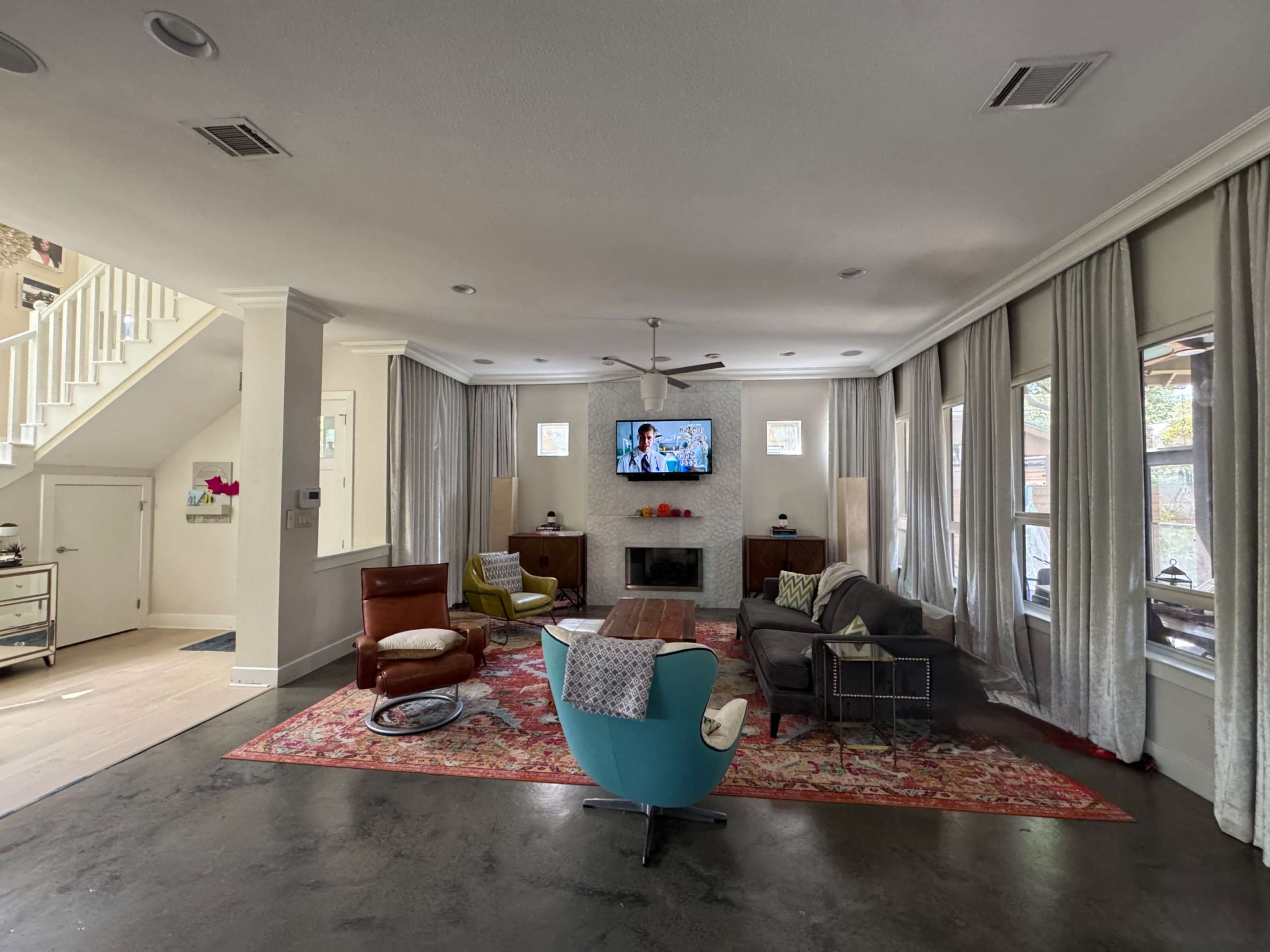 The image shows a modern living room with a television mounted on the wall, a mix of seating options including colorful chairs and a brown leather sofa, and large windows draped with light curtains.