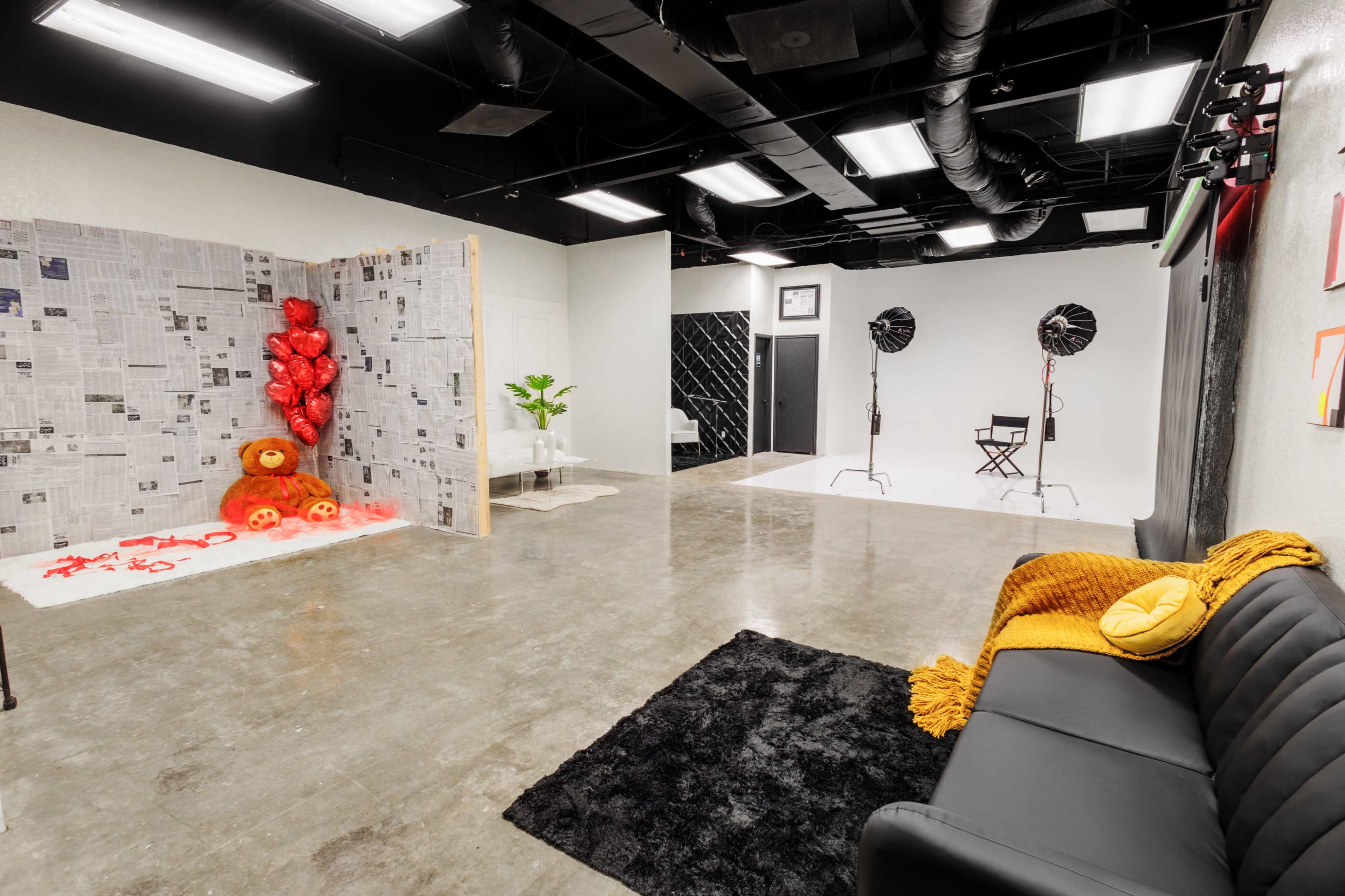 A photography studio featuring a black sofa, a section with newspaper-covered walls and red heart balloons, and a backdrop area with lighting equipment.