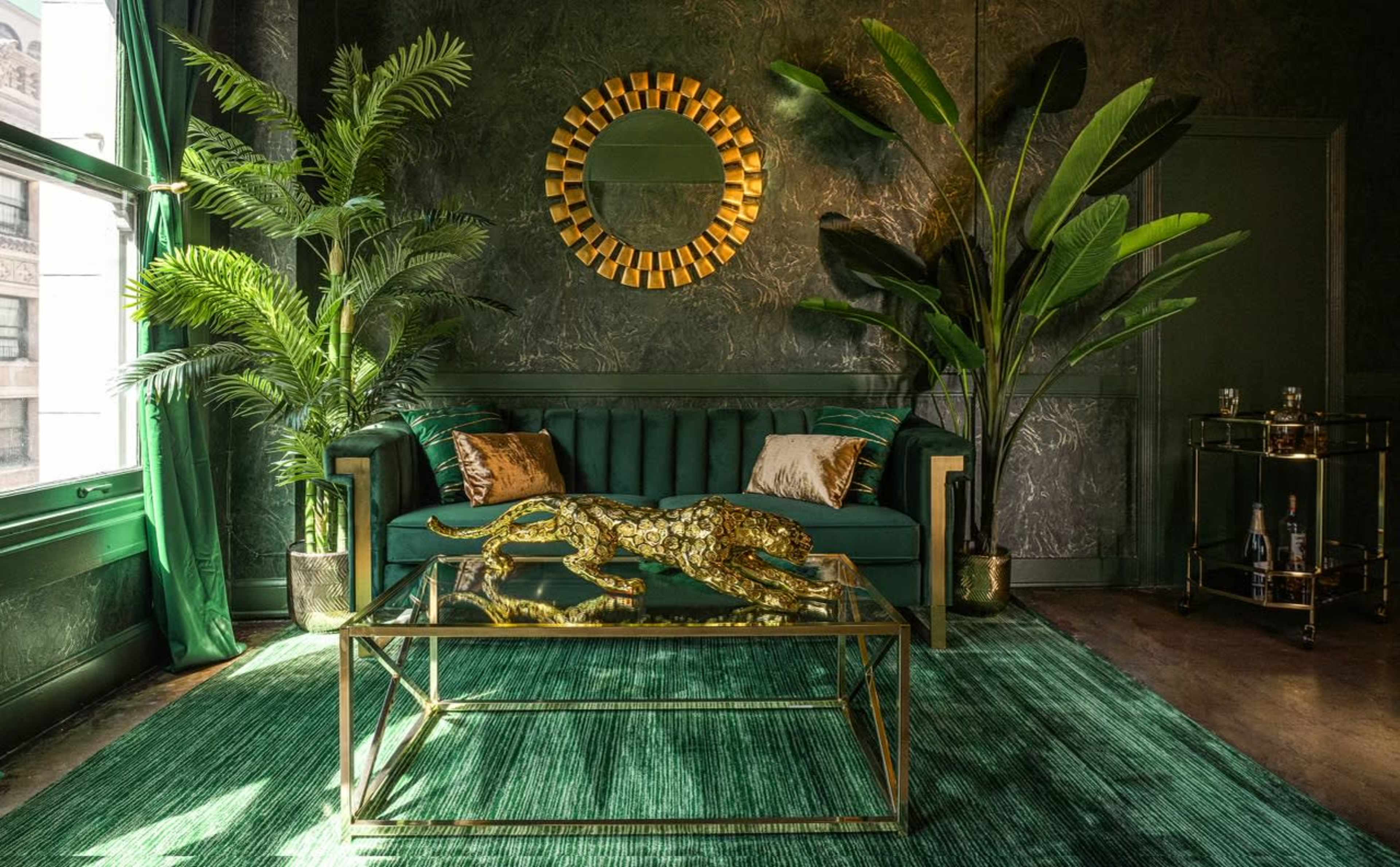 A stylish room featuring a green velvet sofa, a glass coffee table with a decorative golden leopard, and large indoor plants against dark textured walls.