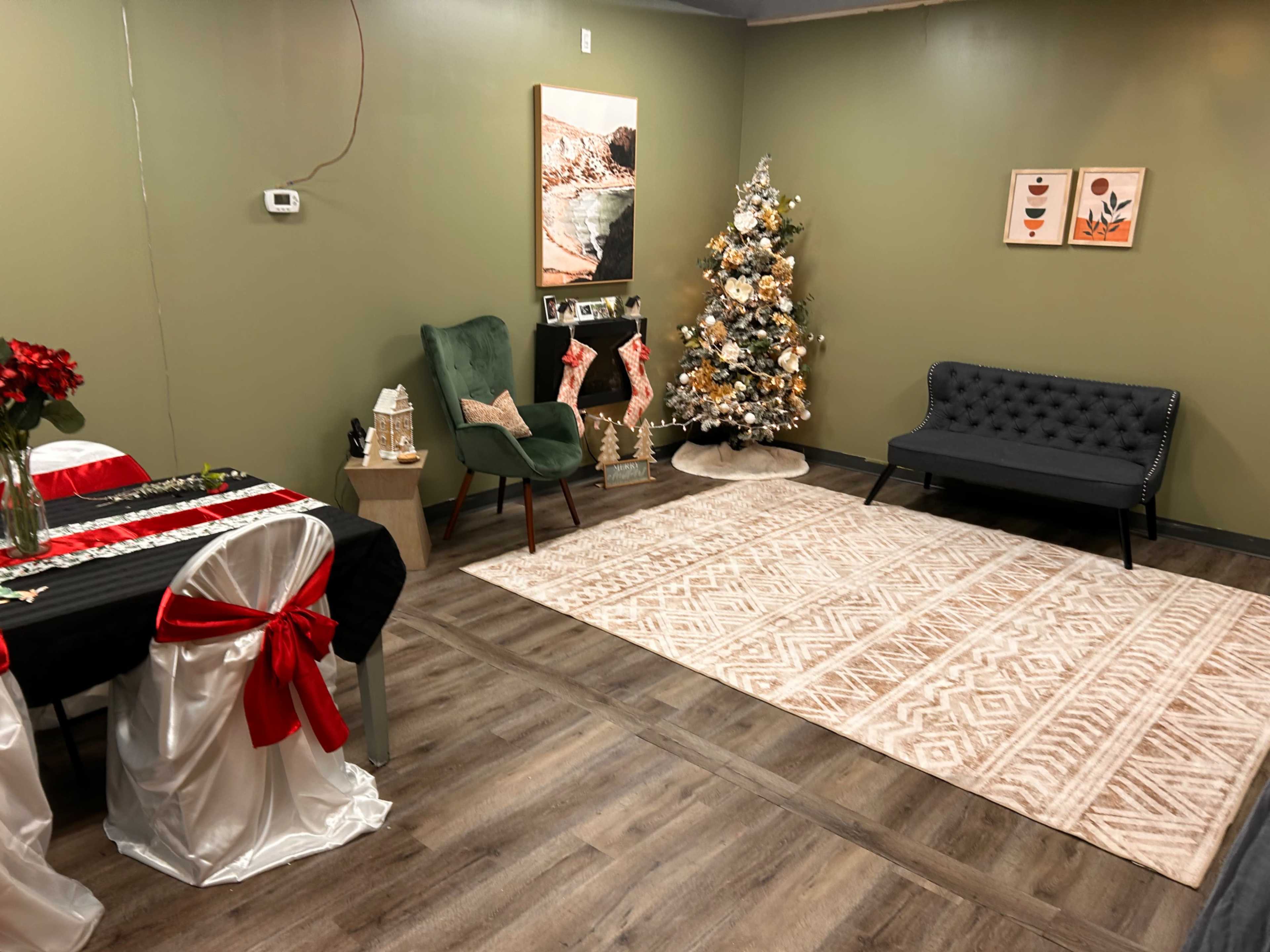 The image shows a cozy indoor space with a decorated Christmas tree, a black sofa, a green chair, a table set with a black and white tablecloth, and a light-colored area rug.