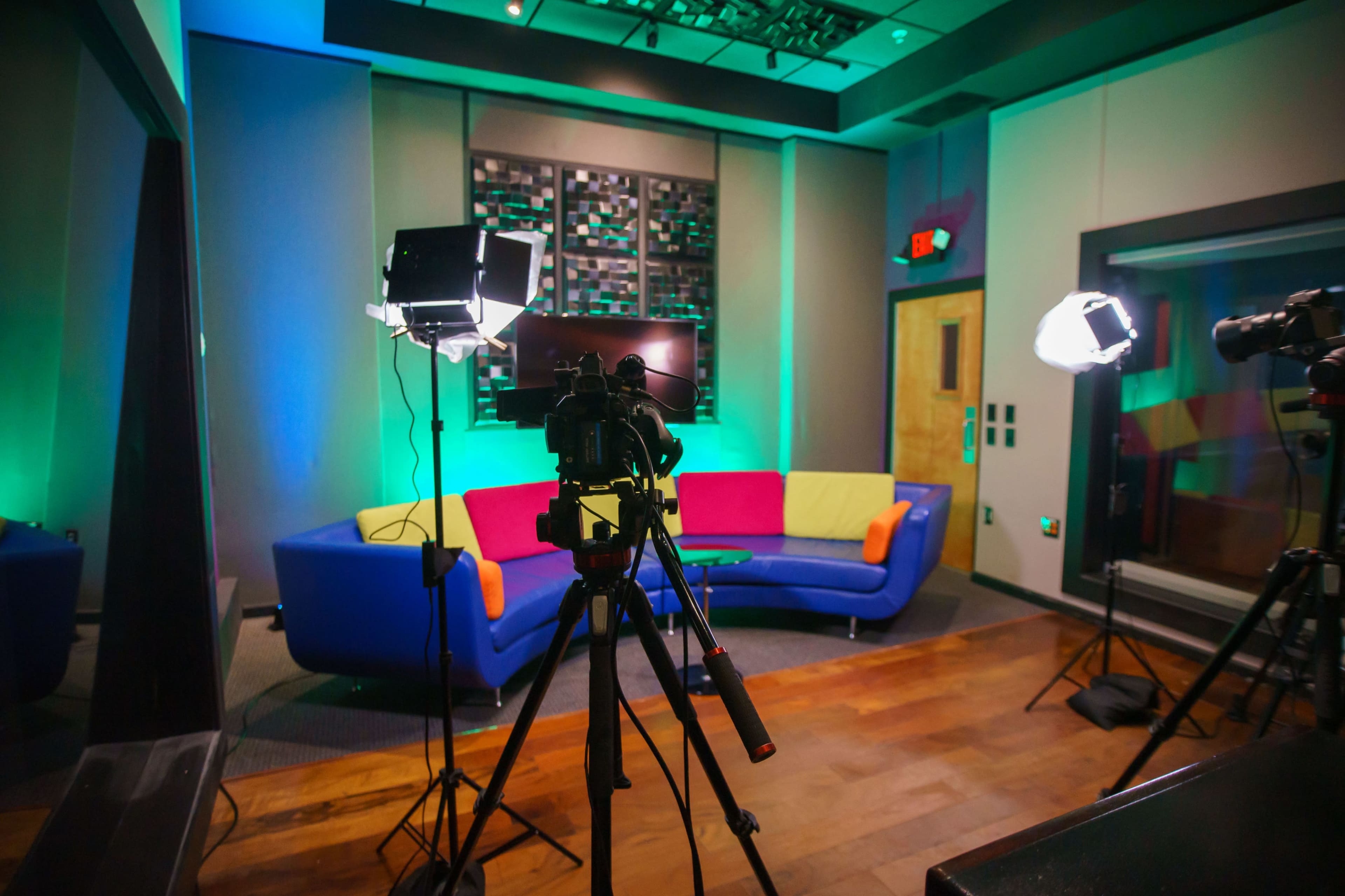 The image shows a brightly lit studio setup featuring a colorful couch, two studio lights, and cameras positioned on tripods.