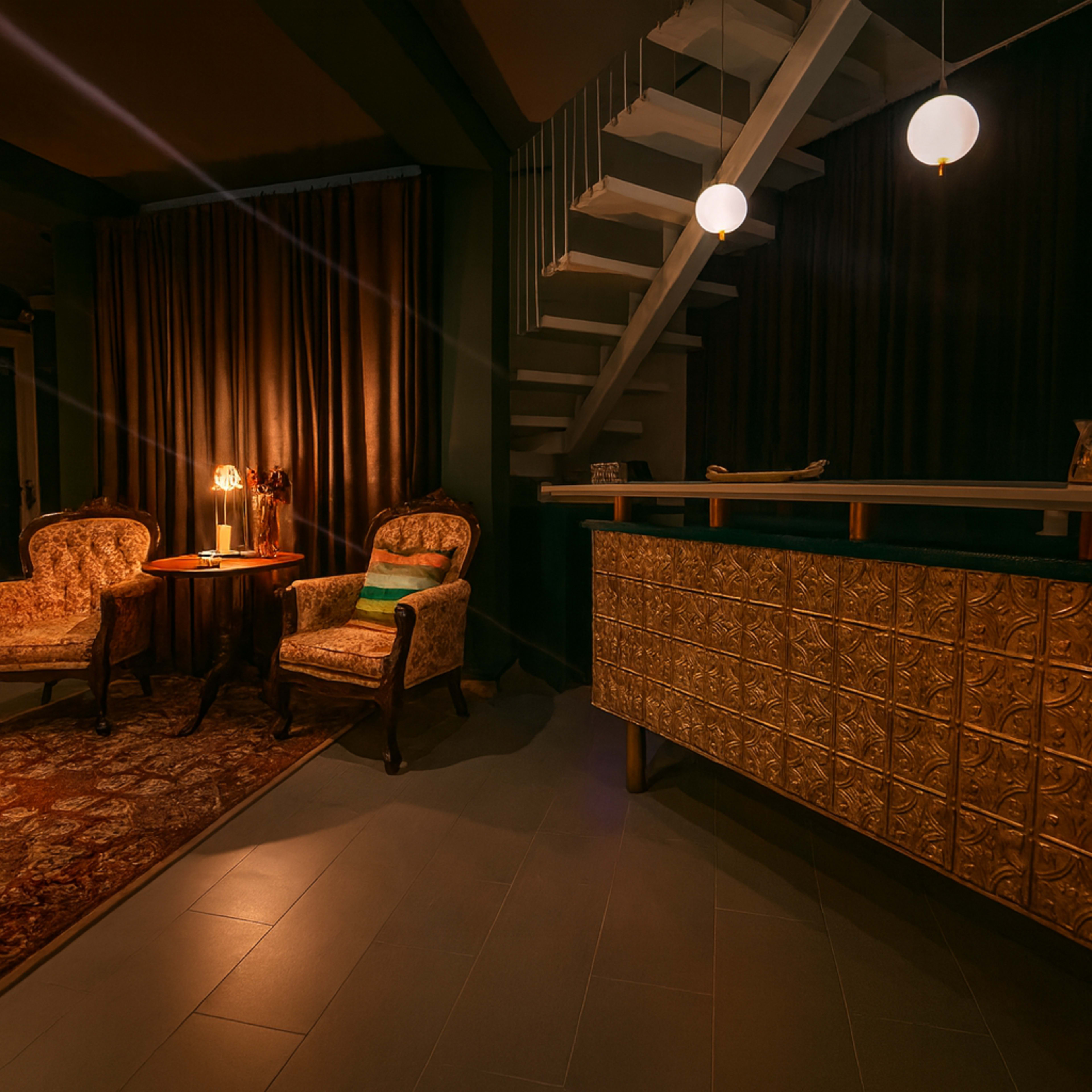 The image shows a cozy interior space with two ornate armchairs, a small side table, and a decorative reception counter, illuminated by soft lighting.