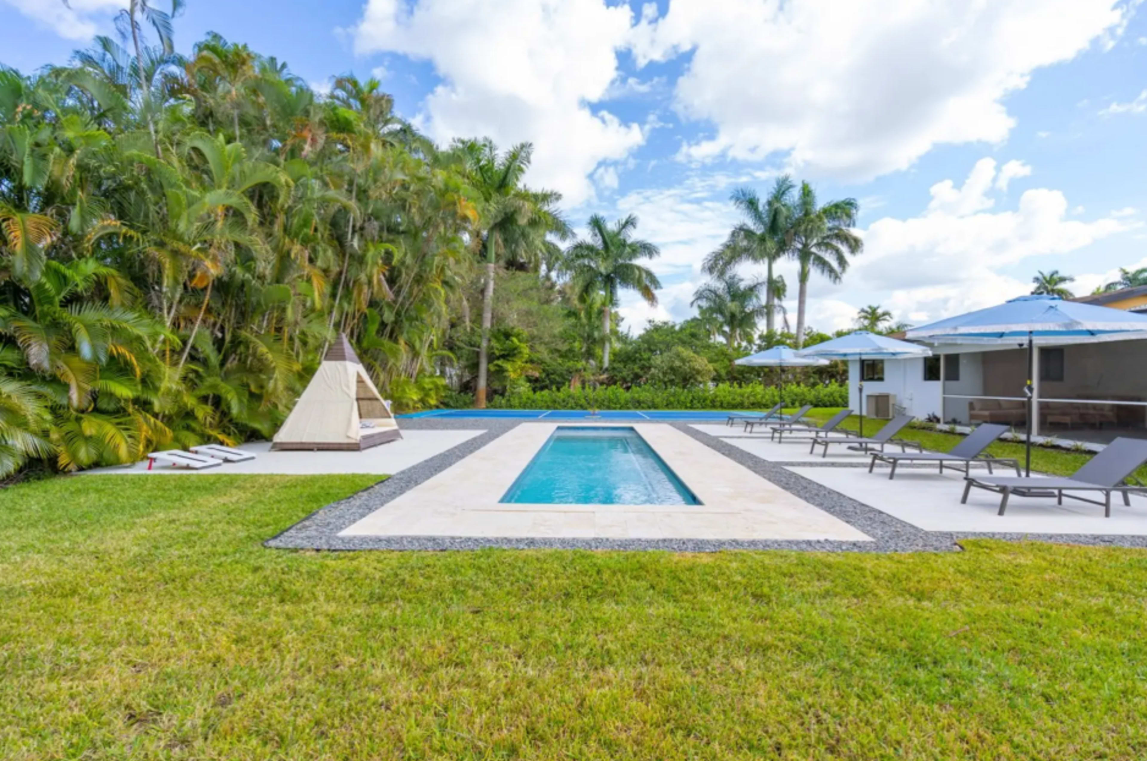 The image shows a swimming pool surrounded by lounge chairs and umbrellas, set in a manicured lawn with tropical plants in the background.