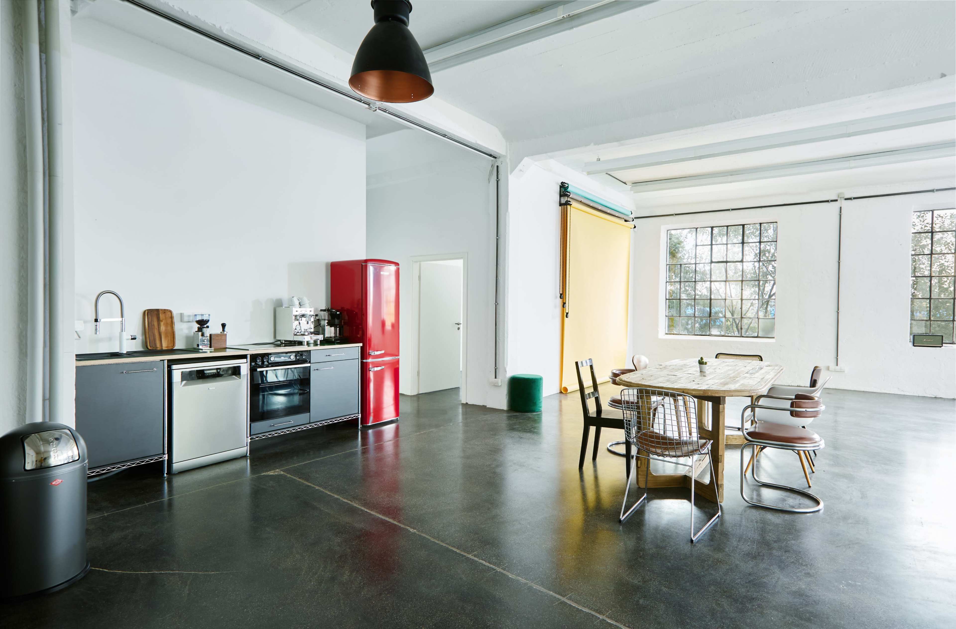 The image shows a spacious kitchen area featuring stainless steel appliances, a bright red refrigerator, a wooden dining table with metal chairs, and large windows providing natural light.