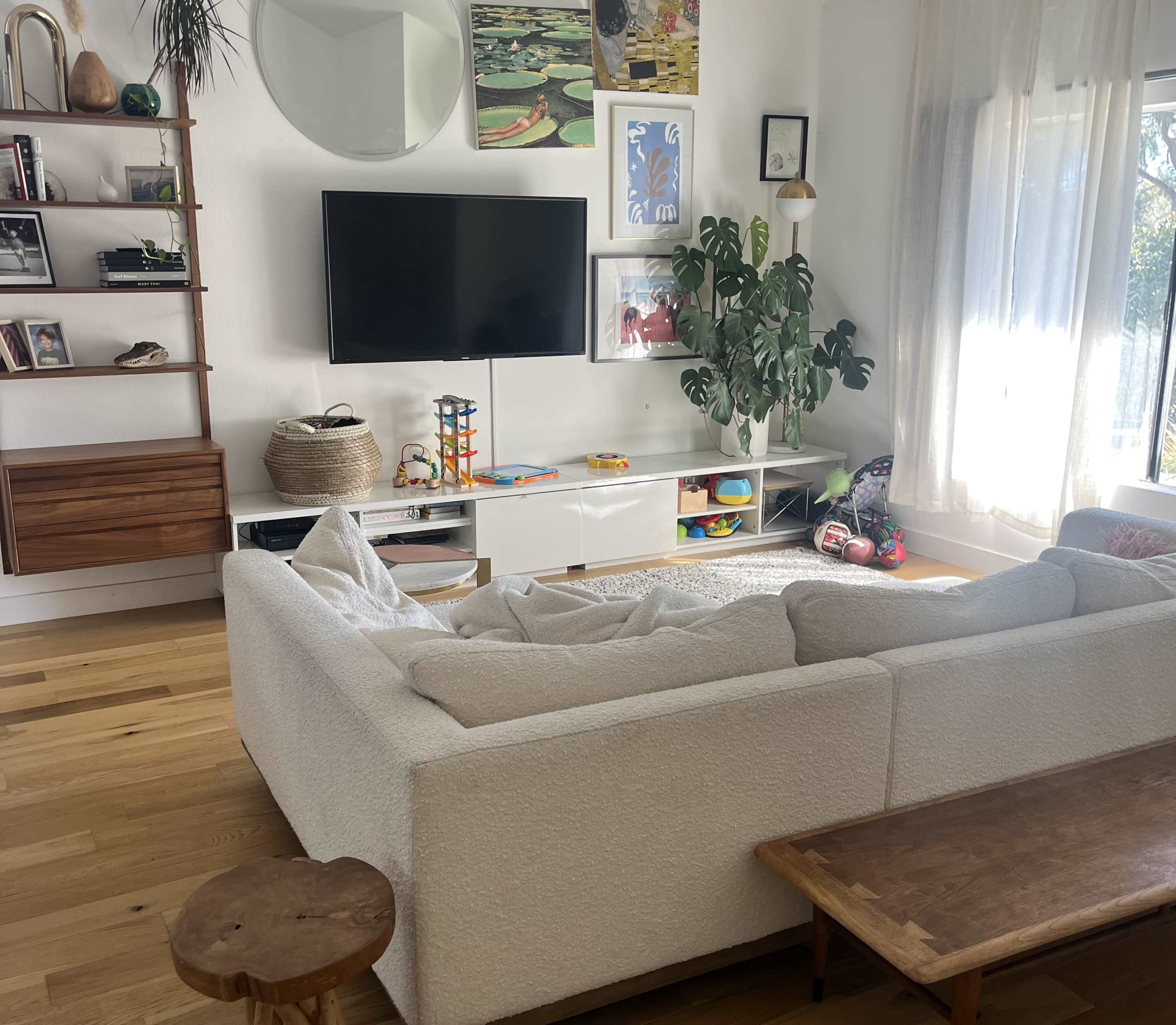 A modern living room features a large, light-colored sofa, a wooden coffee table, a television mounted on the wall, and a variety of decorative items on shelves.