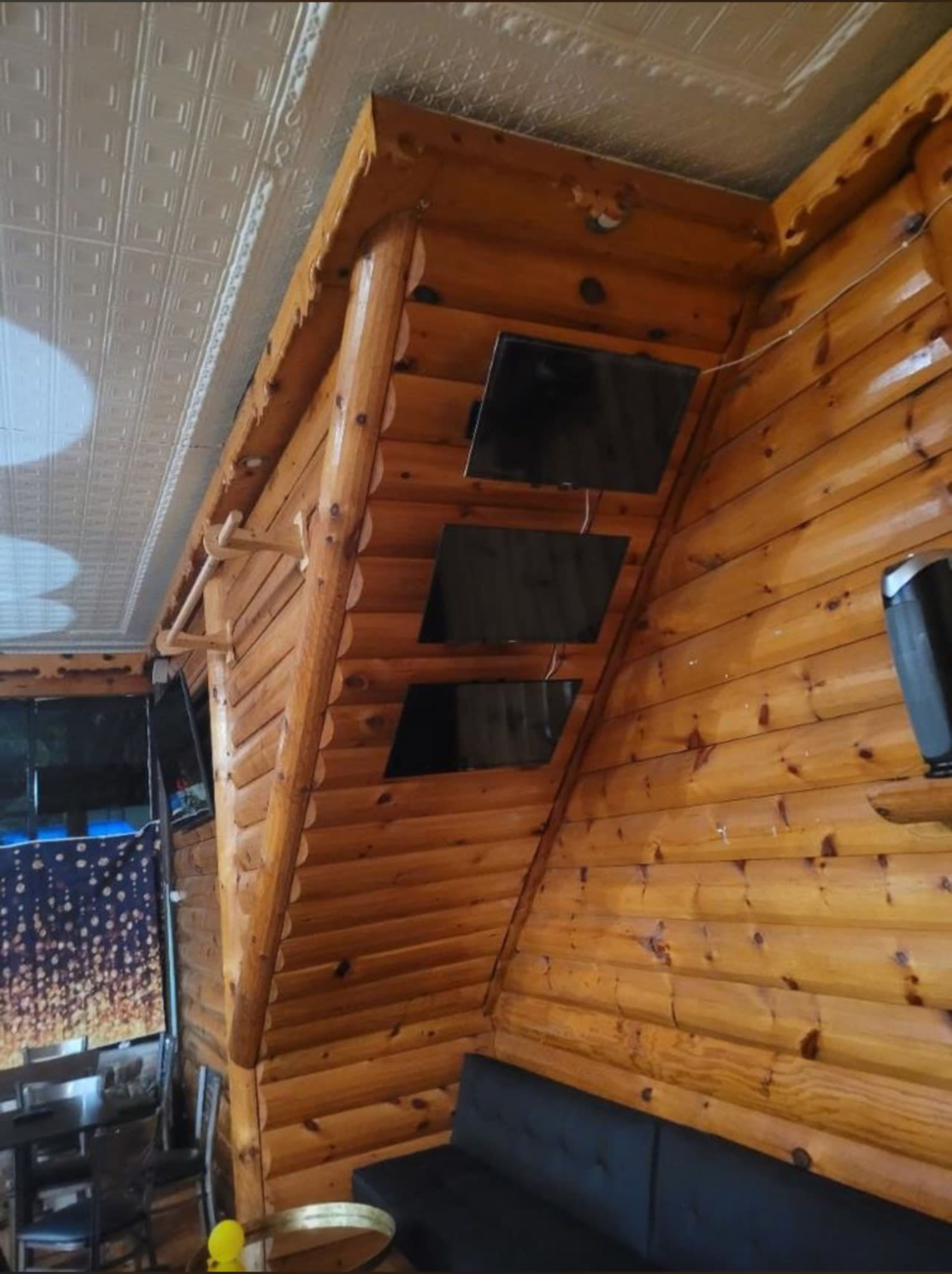 The interior of a wooden cabin with a slanted wall featuring three flat-screen televisions mounted on it.