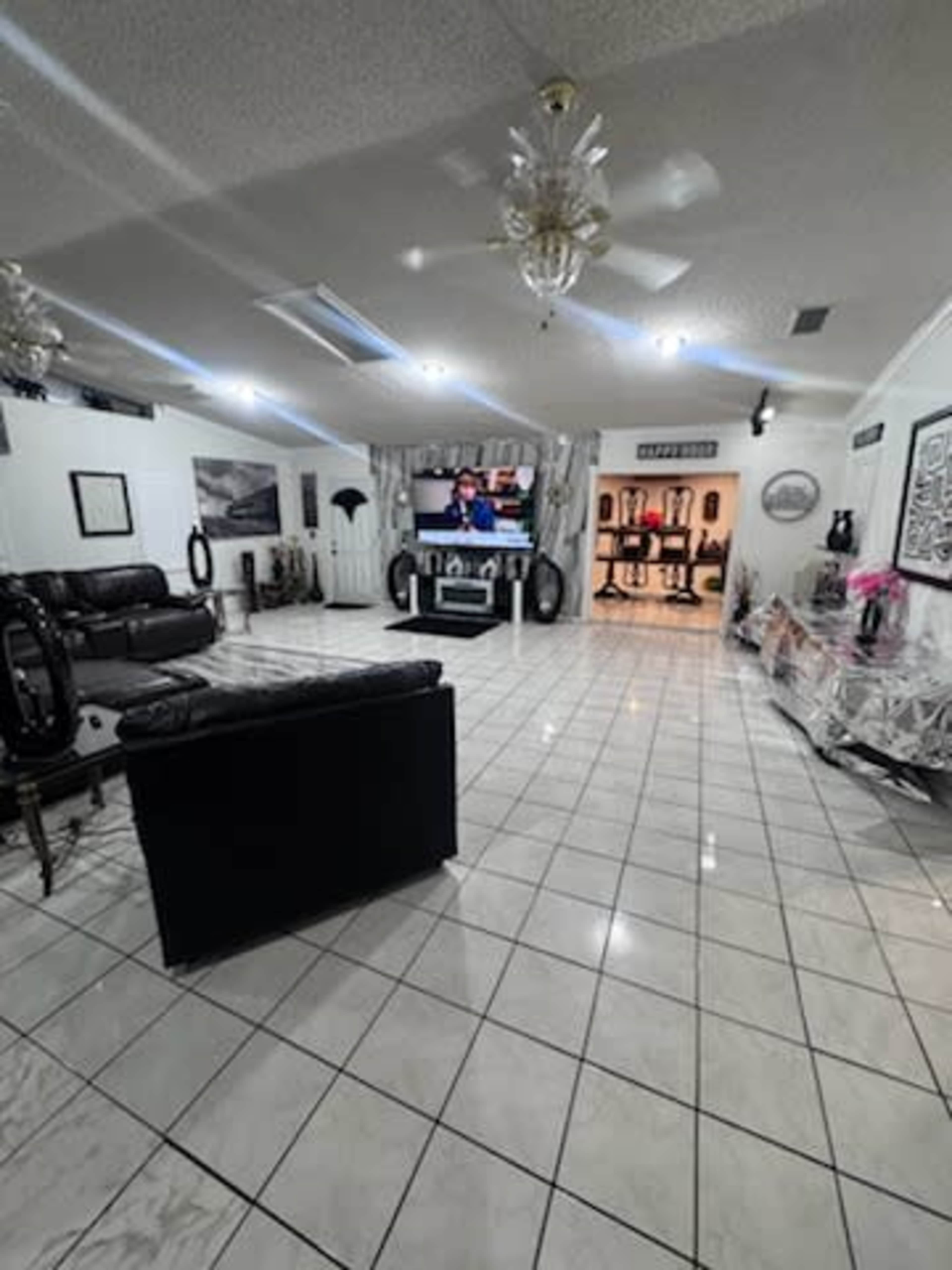 The image shows a spacious living room with black leather sofas, a television mounted on the wall, and a tiled floor reflecting overhead lights.