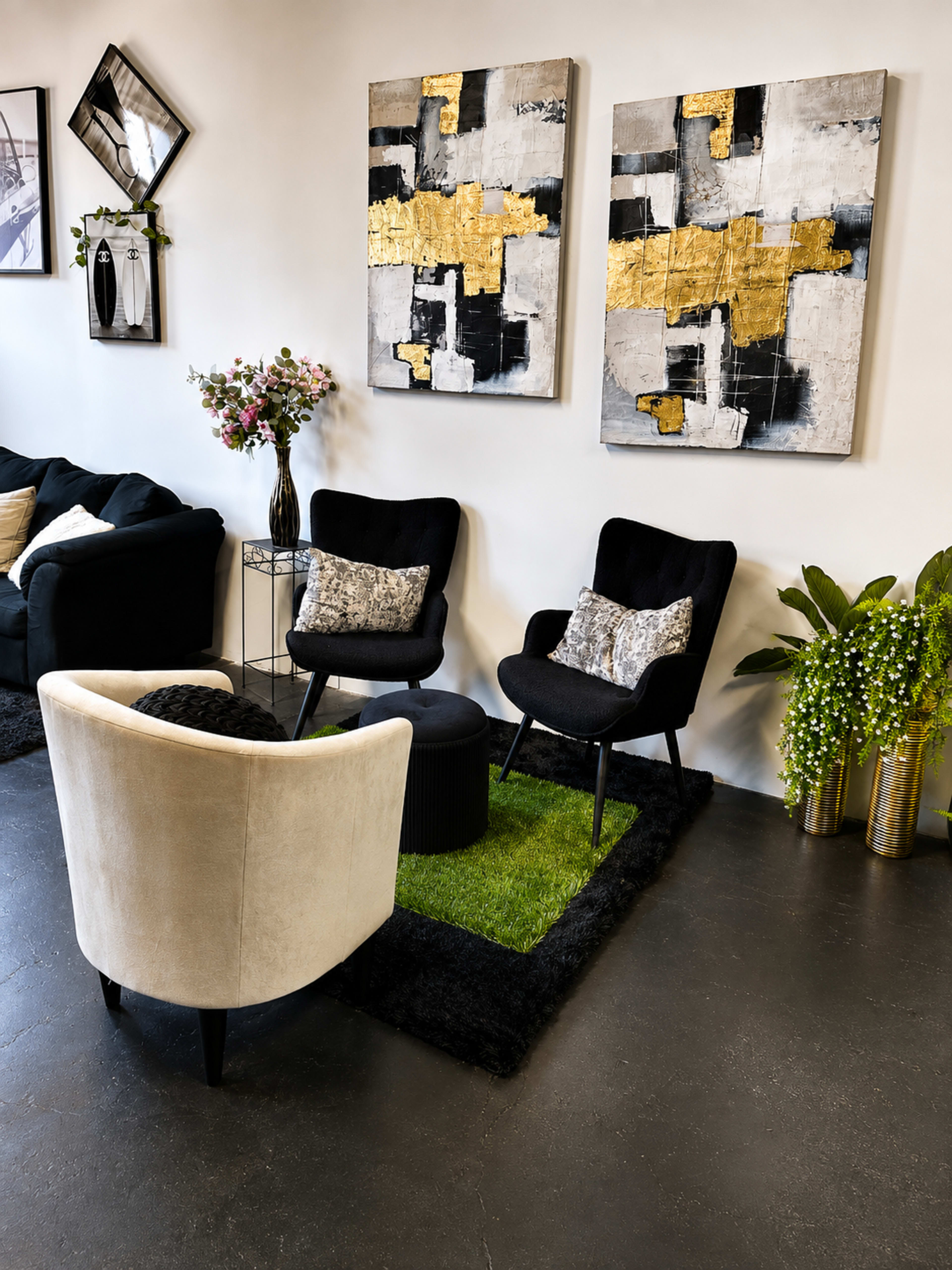The image shows a modern seating area with two black chairs, a white chair, a round black table, and abstract art on the wall, surrounded by plants and floral arrangements.