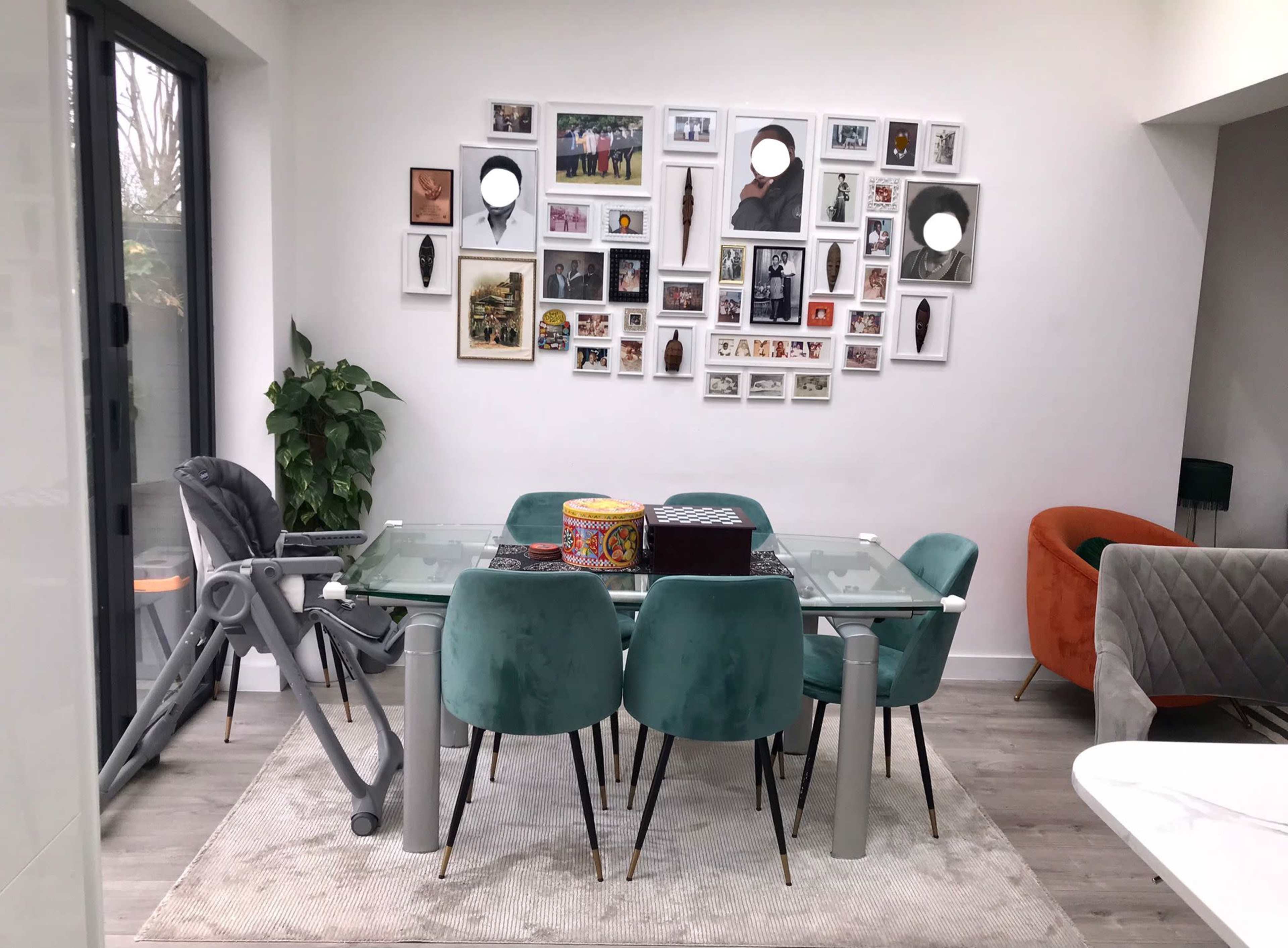 A modern dining area features a glass table surrounded by teal chairs, with a wall displaying a collage of framed photographs and artwork.