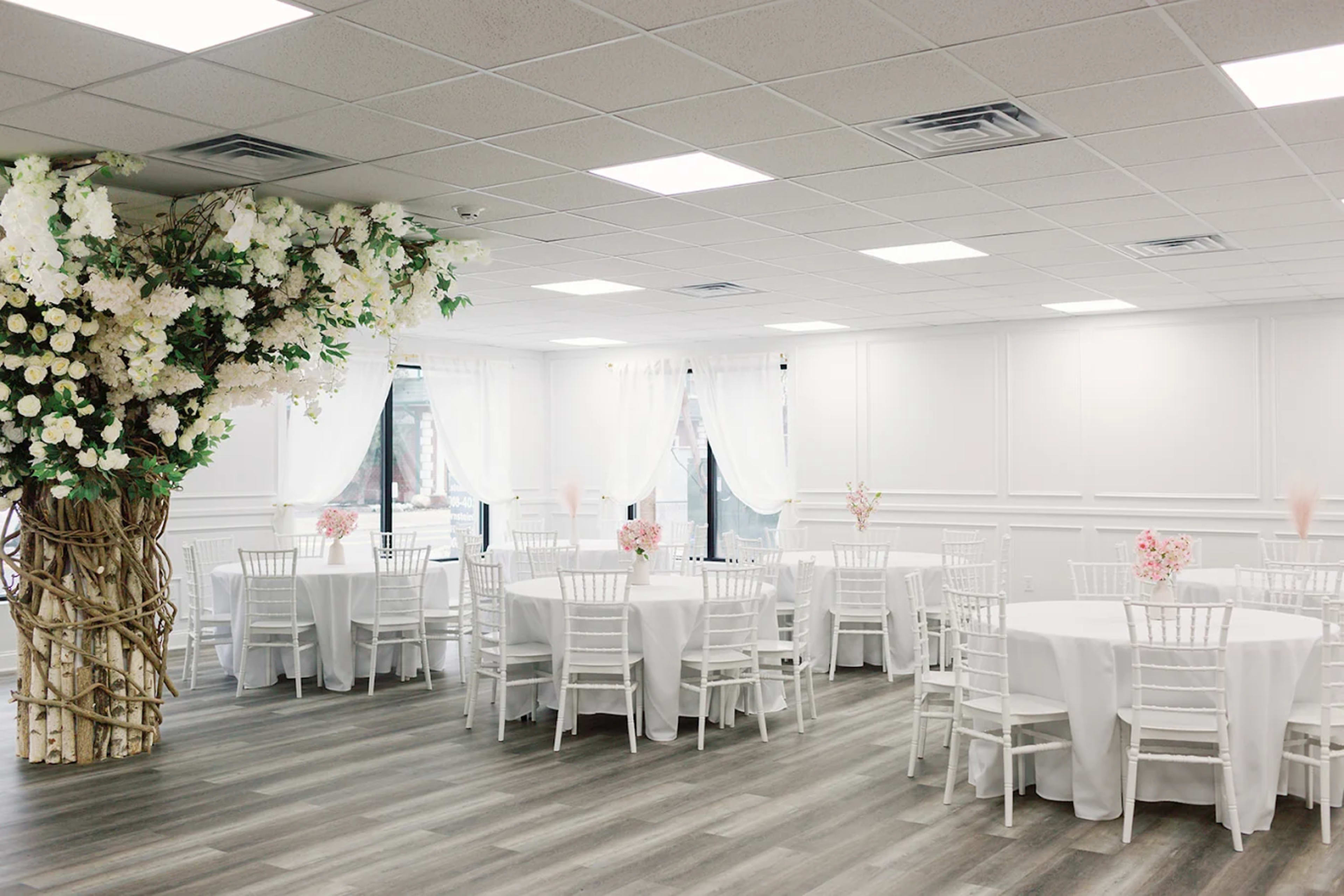 The image shows a spacious event room with white tables and chairs arranged neatly, adorned with floral decorations and sheer white drapes.