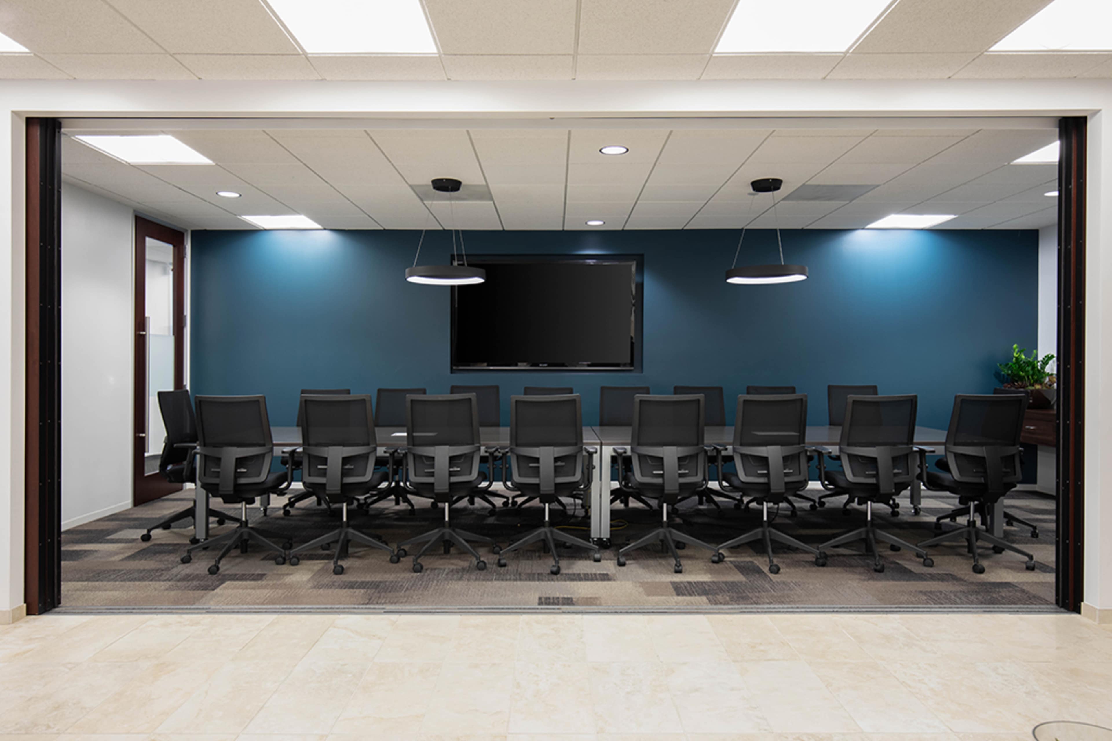A modern conference room features a long table surrounded by ergonomic black office chairs, with a large screen mounted on a teal wall.