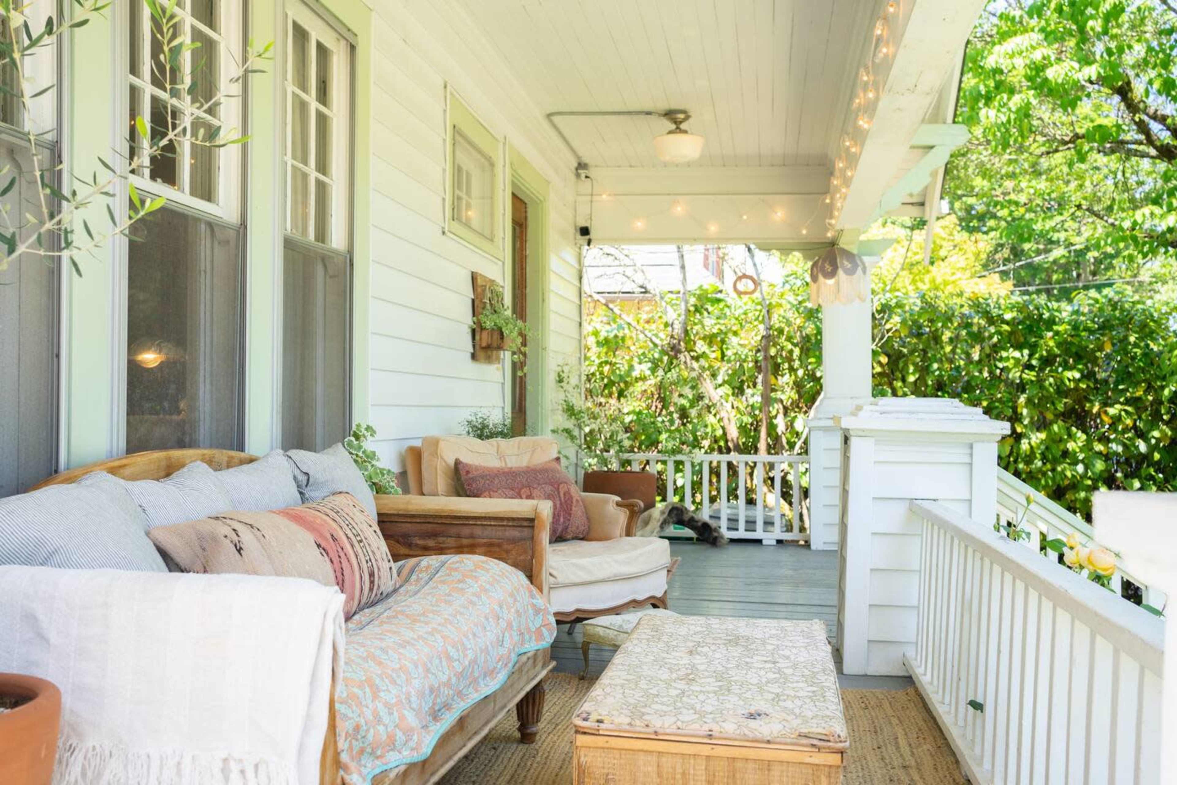 A wooden porch features comfortable seating with cushions, a wicker table, and greenery in the background.