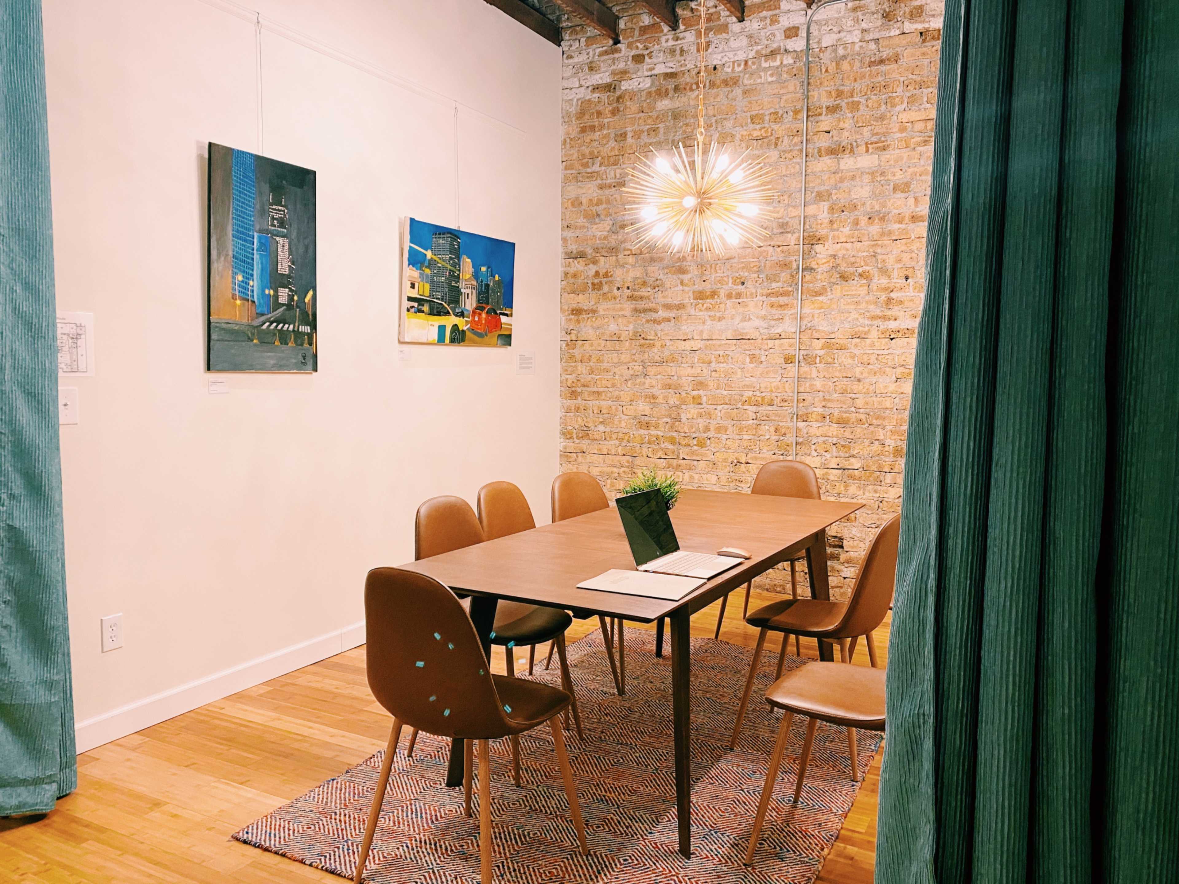 A modern meeting room features a large wooden table surrounded by brown chairs, with artwork on the walls and a pendant light above.