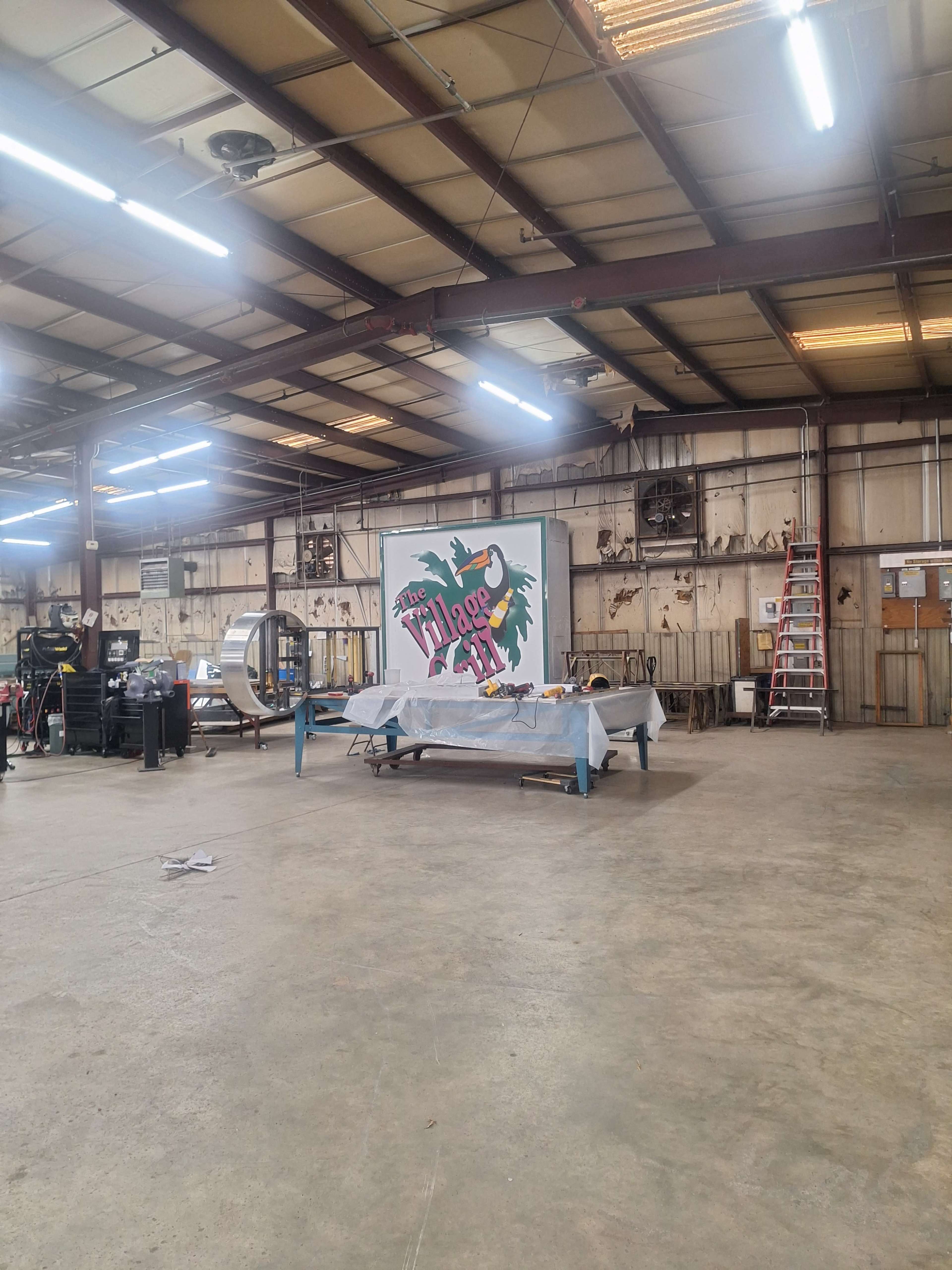 The image depicts a spacious industrial workshop with a large mural on the wall, various tools and equipment scattered throughout, and a ladder leaning against the wall.