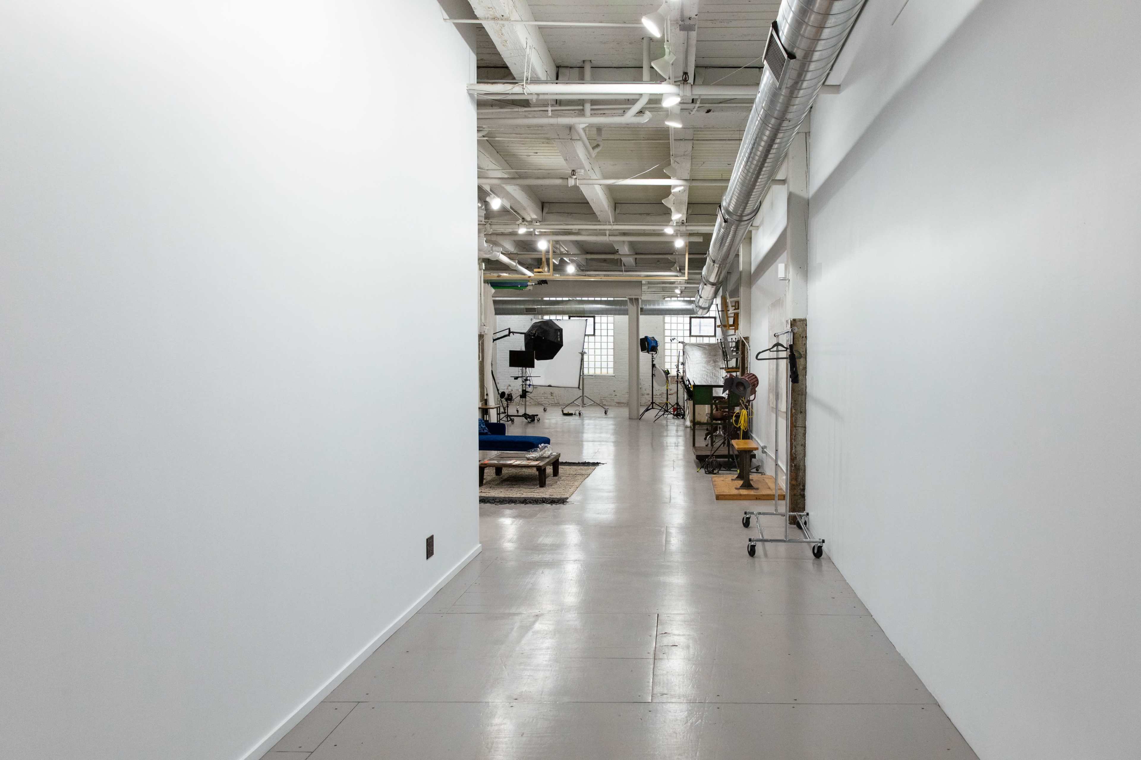 The image shows a long, brightly lit corridor leading to a spacious studio with photography equipment and props.