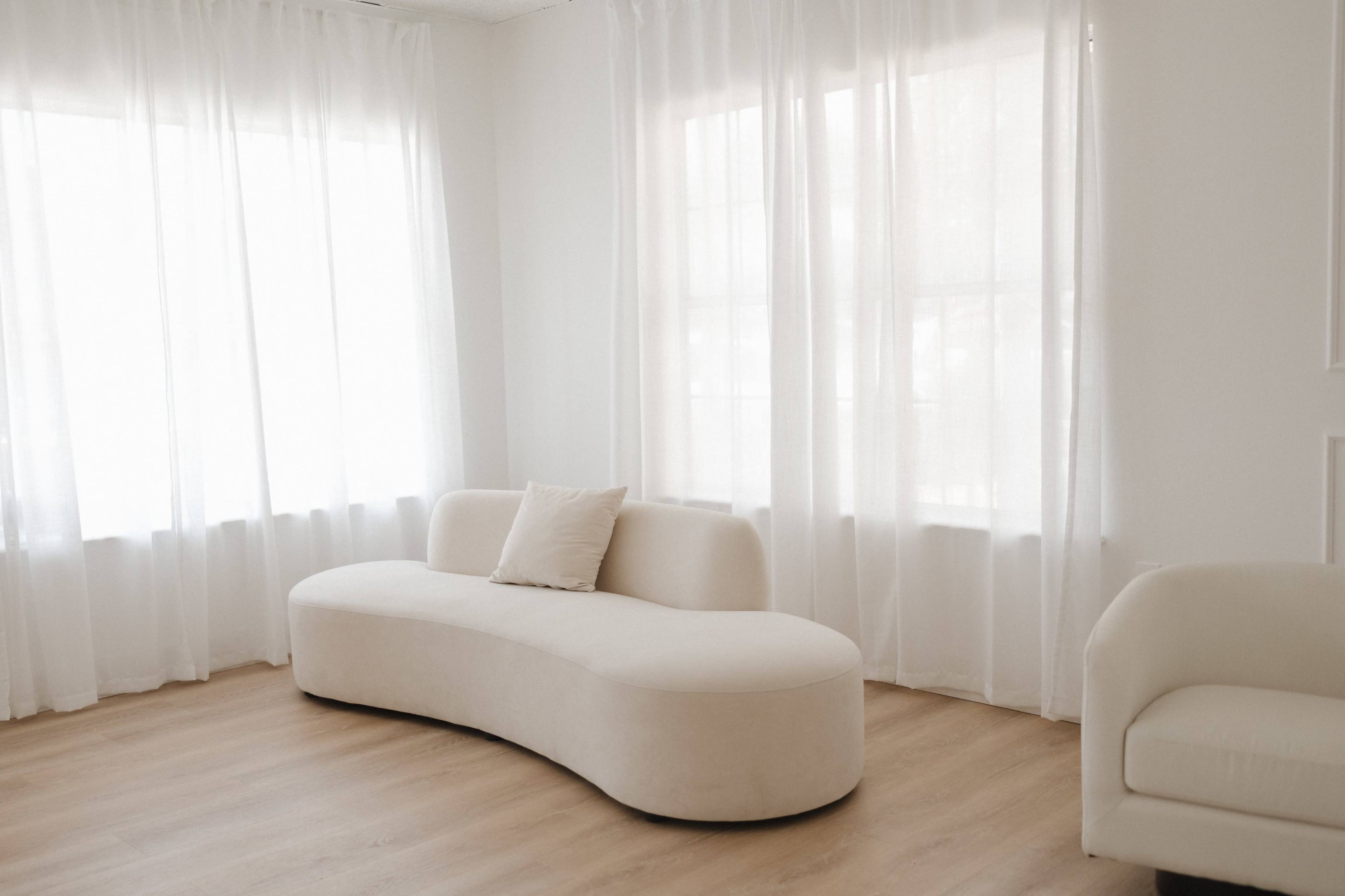 A curved beige sofa is positioned against a wall with large windows covered by white curtains in a softly lit room with wooden flooring.