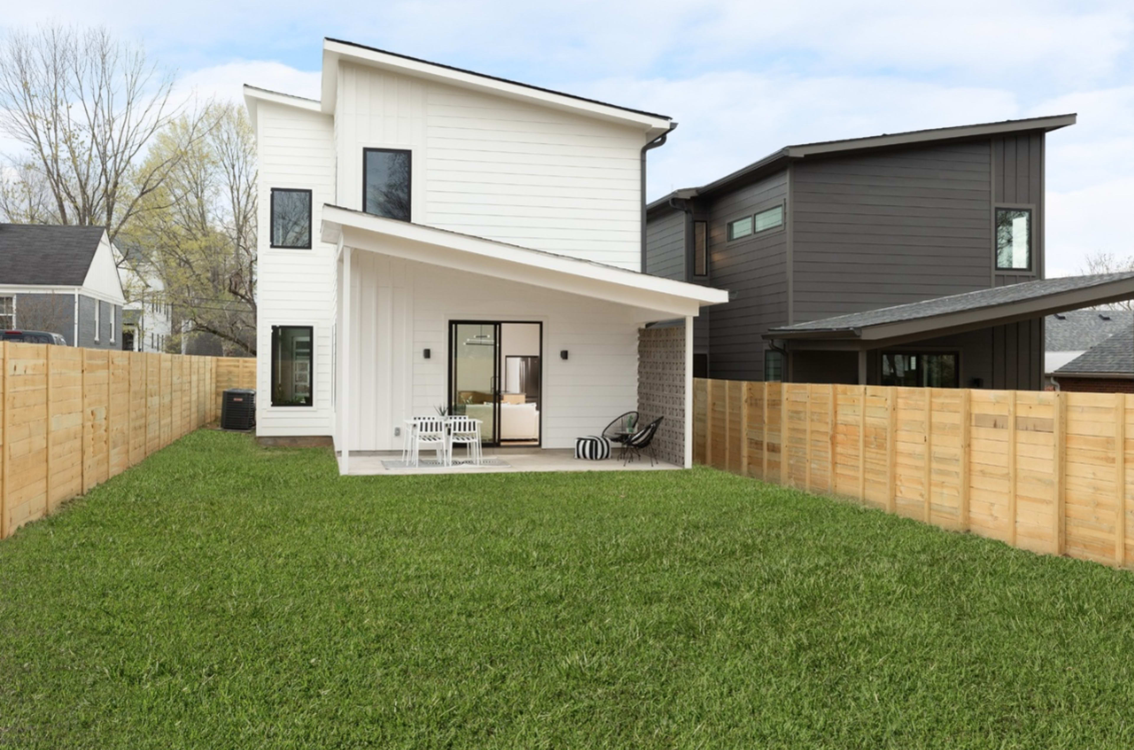 The image shows a modern two-story house with a sloped roof, surrounded by a fenced yard featuring green grass.